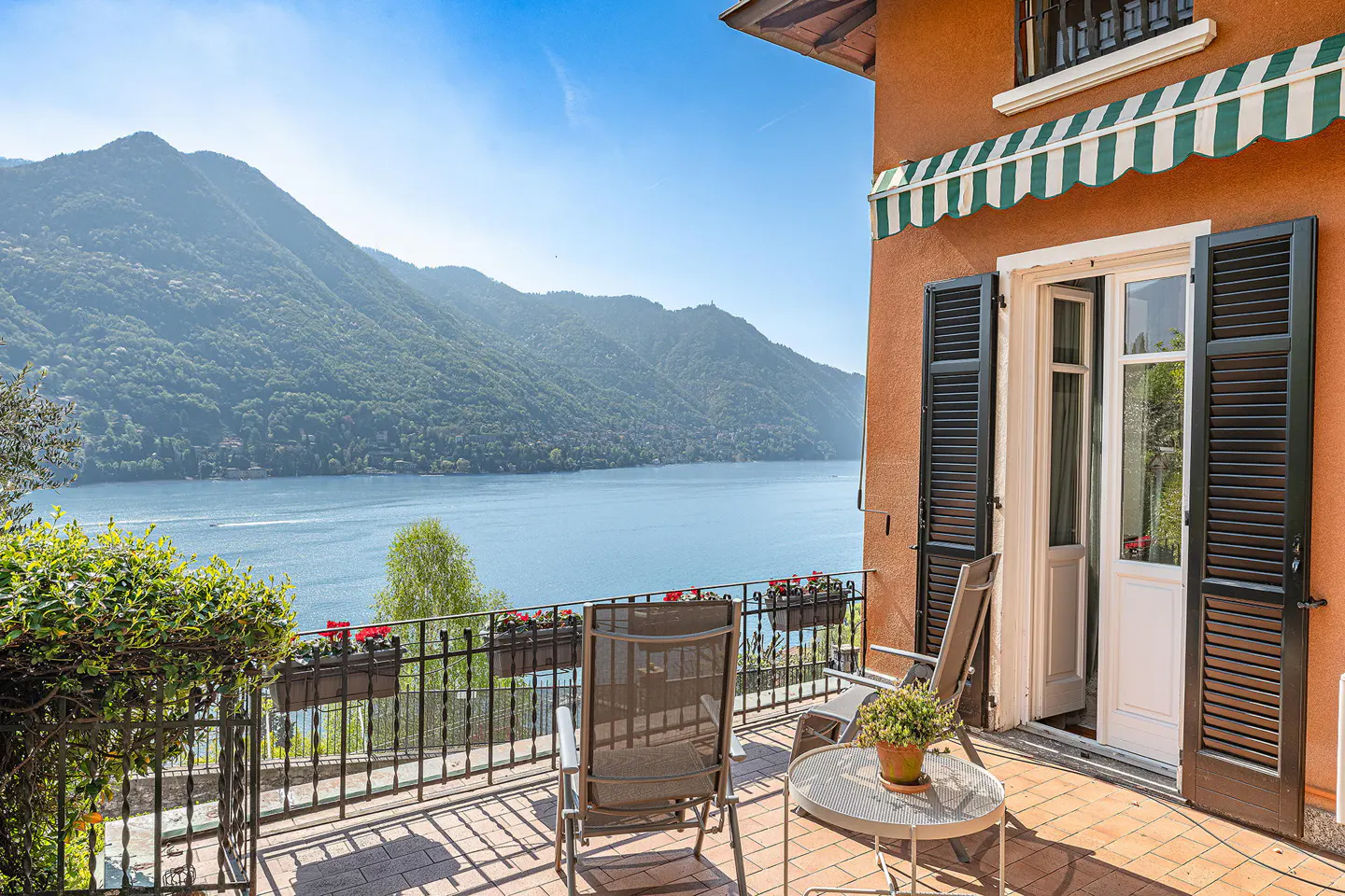 Balcony with chairs and table overlooks Lake Como, Italy. Orange building with green striped awning and black shutters.