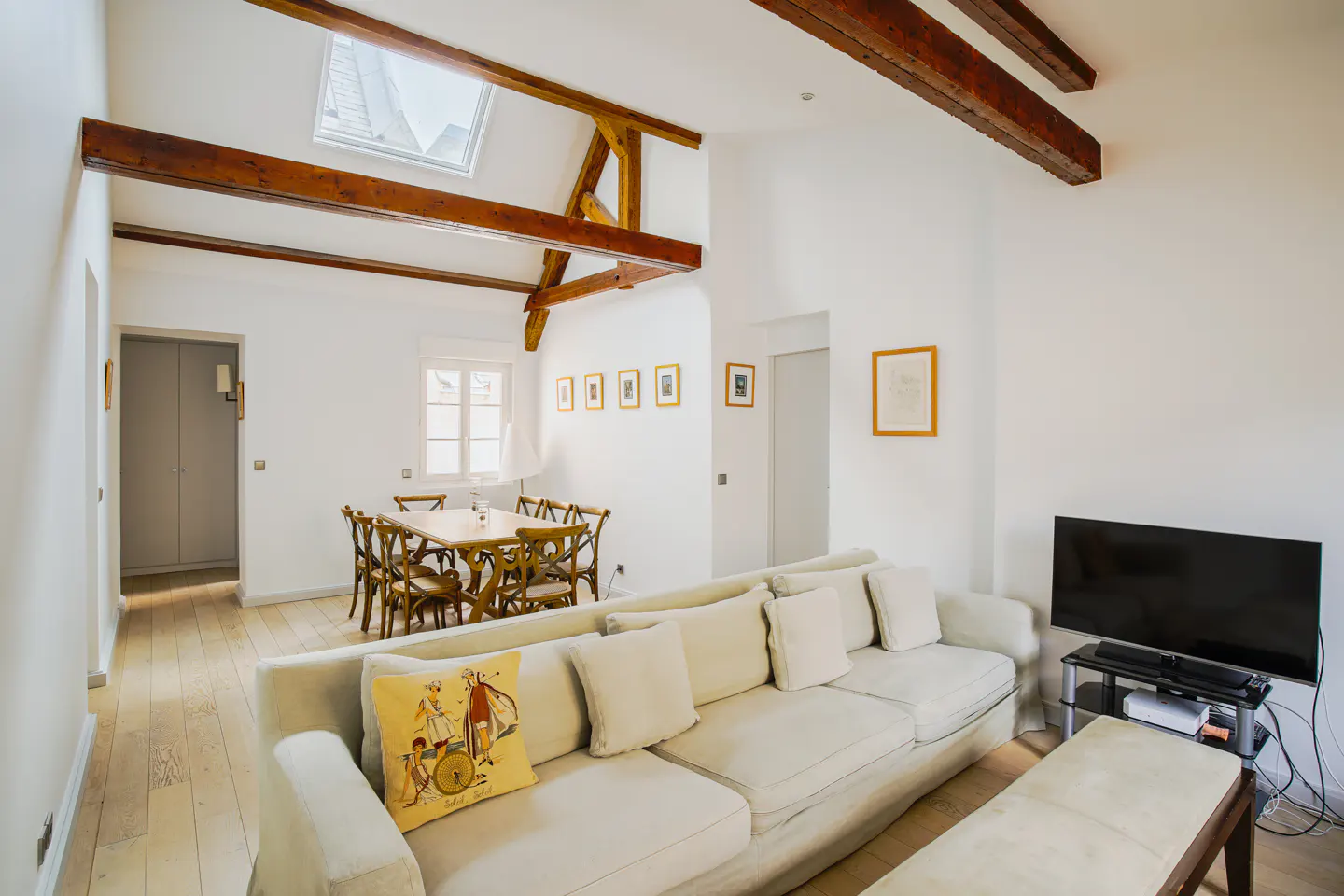 Bright living room with white walls, wooden beams, and light wood floors. A cream sofa faces a TV. A dining table with chairs is in the background.