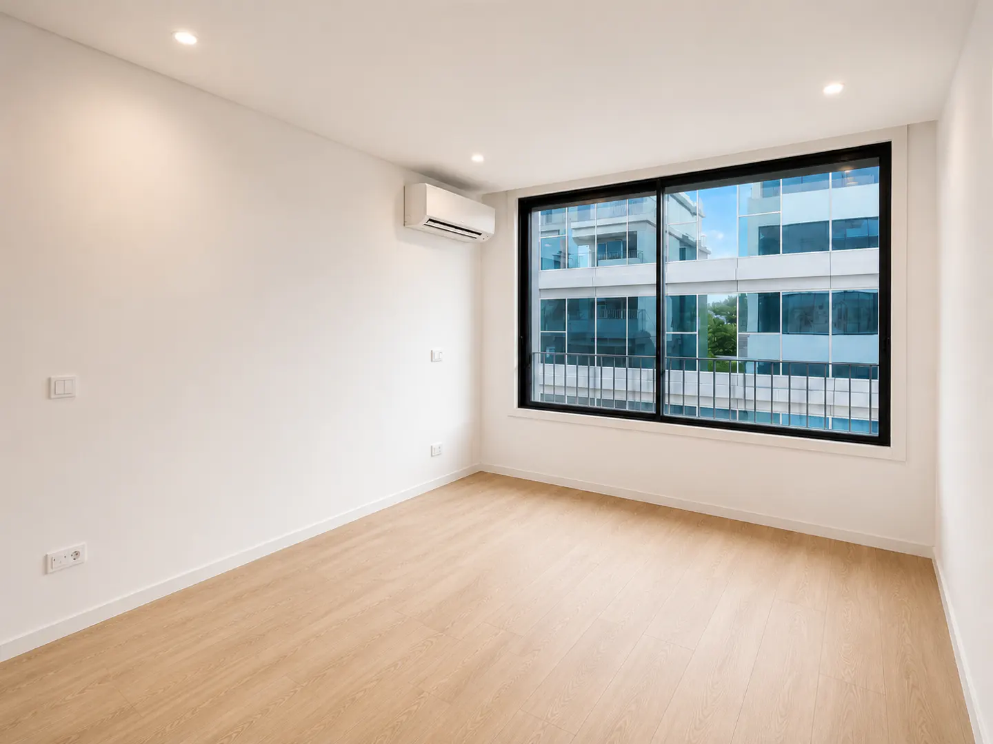 Bright, empty room with light wood floors, white walls, and a large window with a black frame showing a building outside.