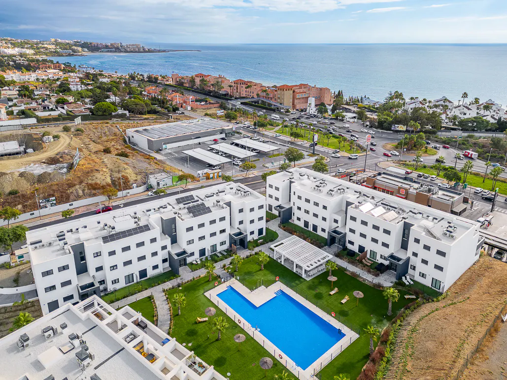 Aerial view of white apartment buildings with a blue pool, green lawn, and ocean backdrop.