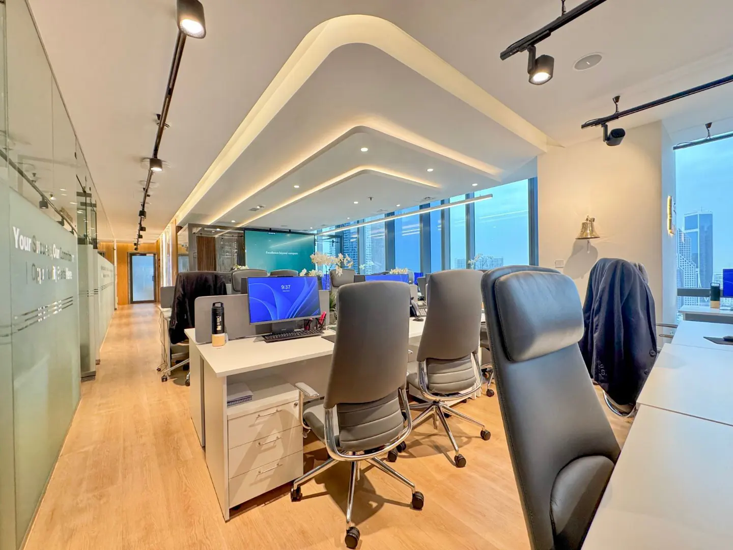 Bright, modern office space with white desks, gray chairs, and wood floors. Large windows offer a city view.