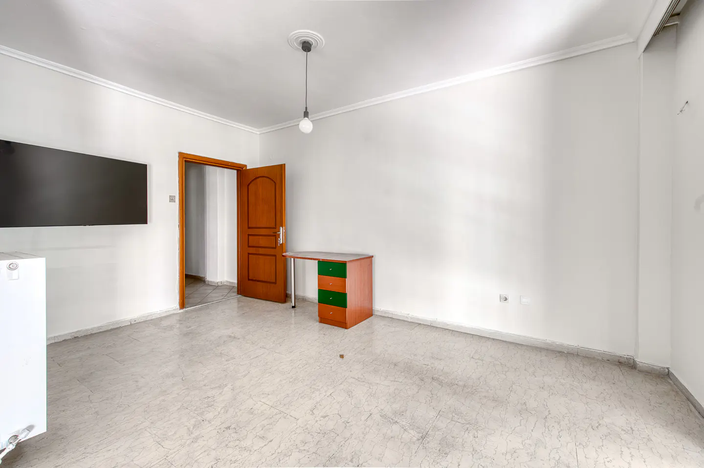 Bright, empty room with marble floors, white walls, and a brown desk with green drawers. A TV hangs on the wall, and a wooden door is ajar.