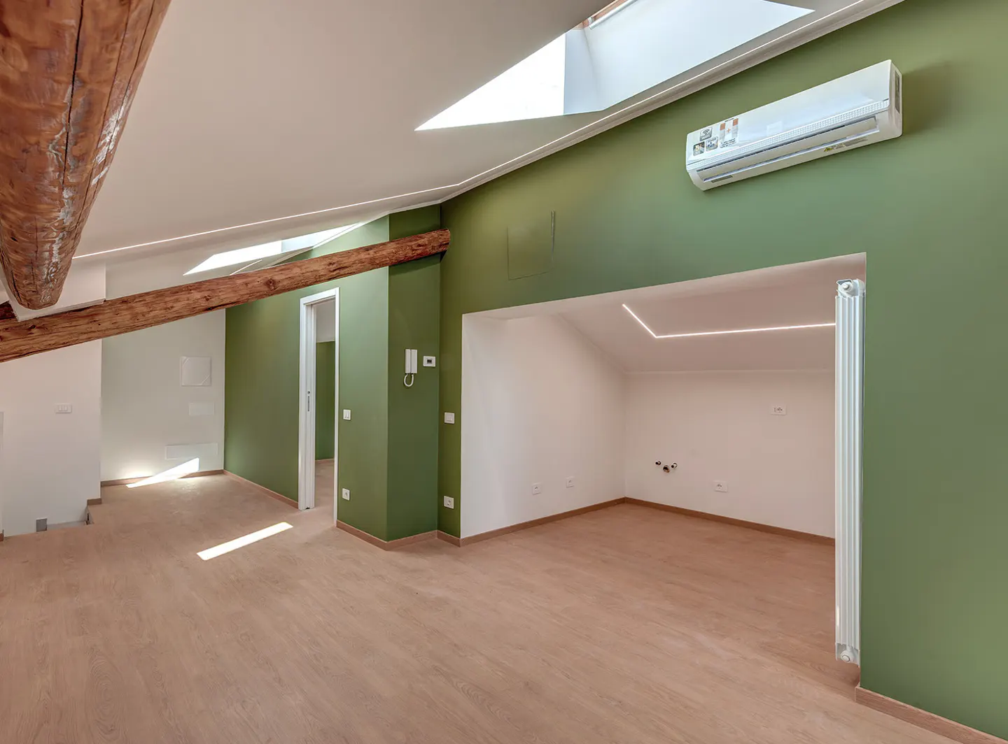 Bright, empty room with wood floors, green accent wall, and exposed wood beams. Skylights provide natural light. Air conditioner on the wall.