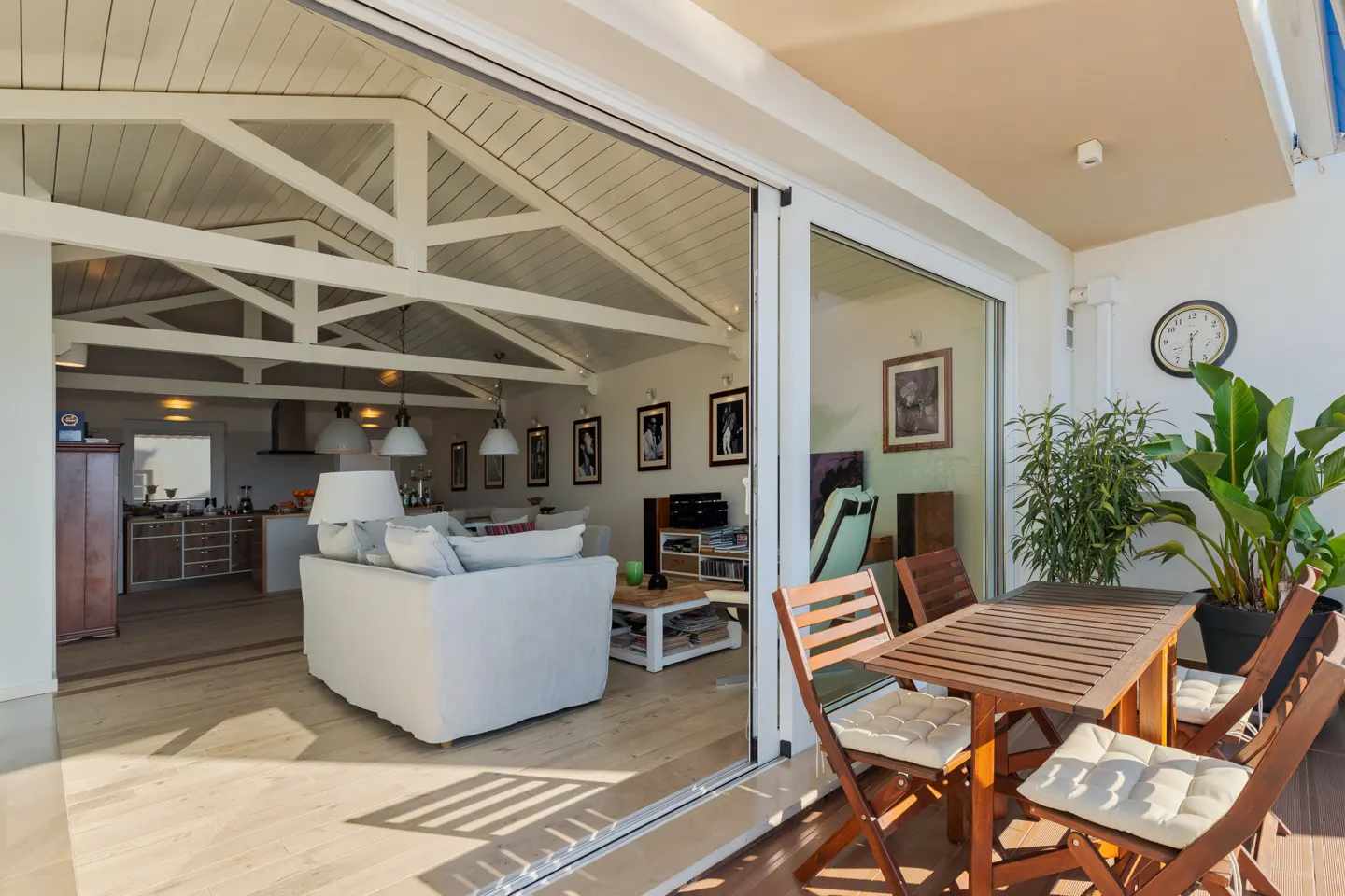 A bright living room with a white sofa opens onto a patio with a wooden dining set and potted plants.