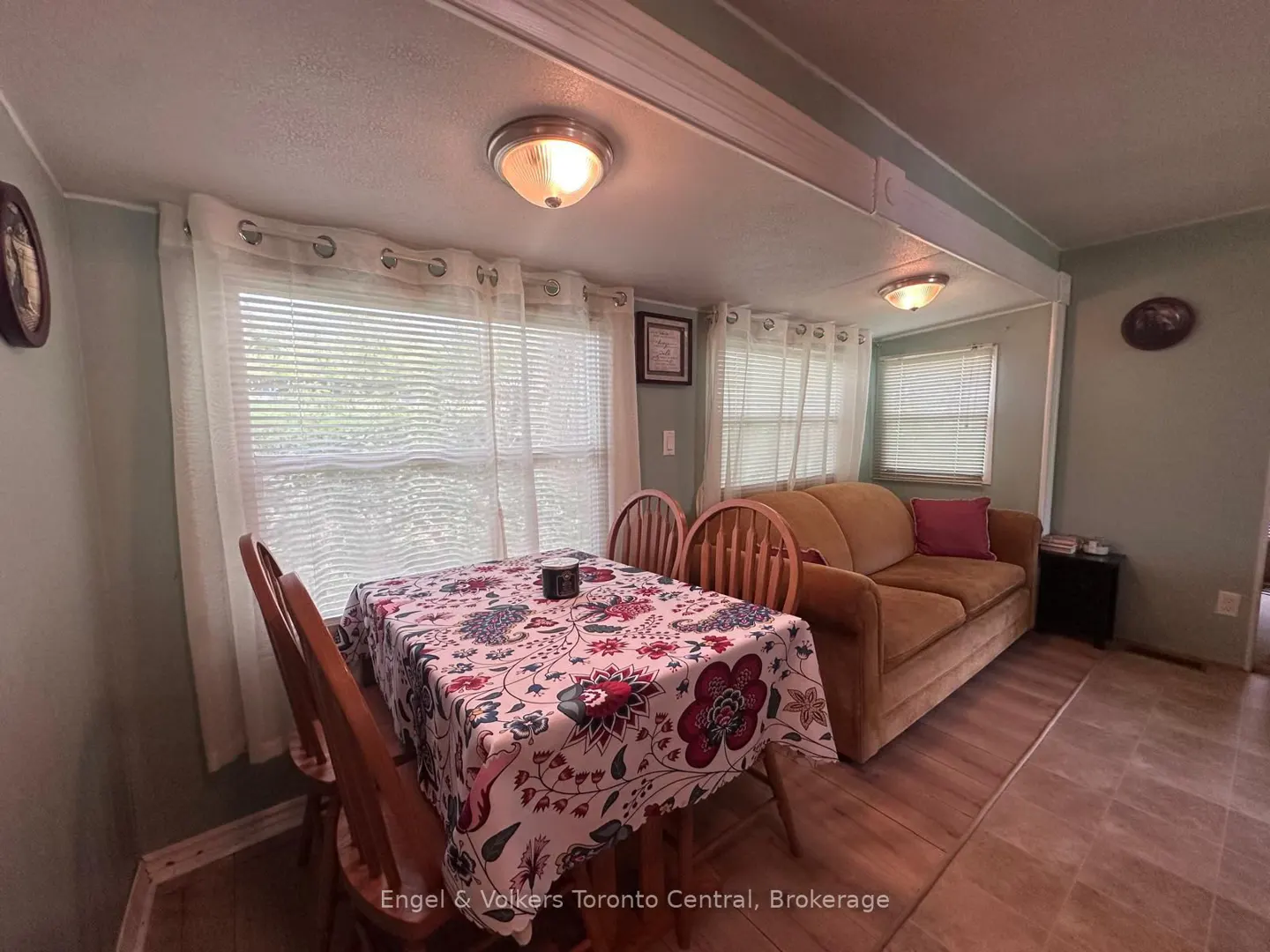 Living room with a floral tablecloth on a wooden table, chairs, and a tan sofa with a pink pillow. Natural light from window.