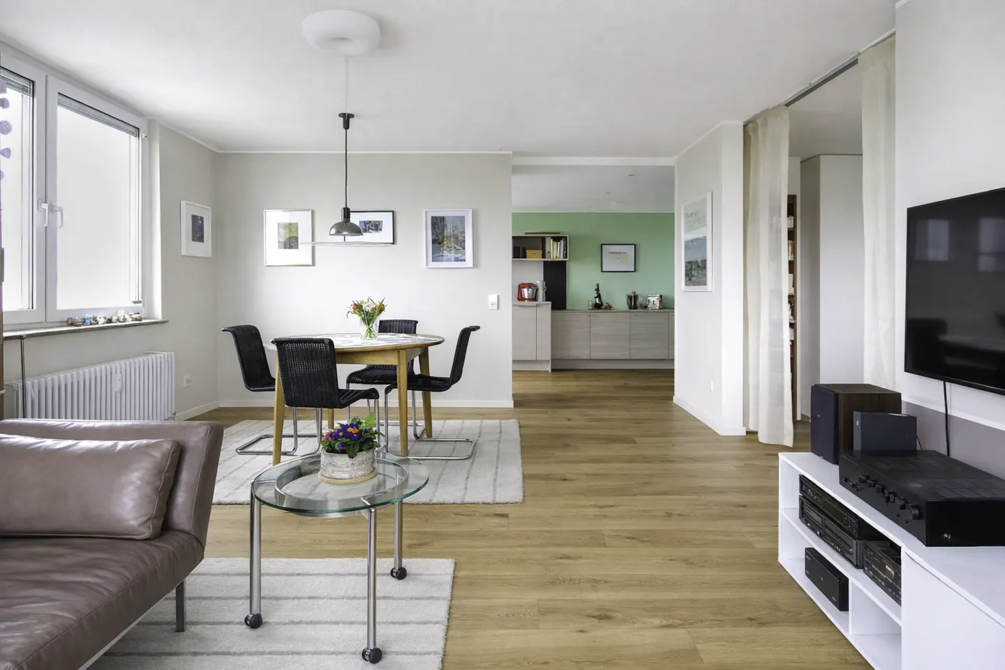 Bright, modern living room with wood floors, a brown leather couch, and a glass coffee table. A dining table with black chairs sits near a window. The kitchen has green walls.