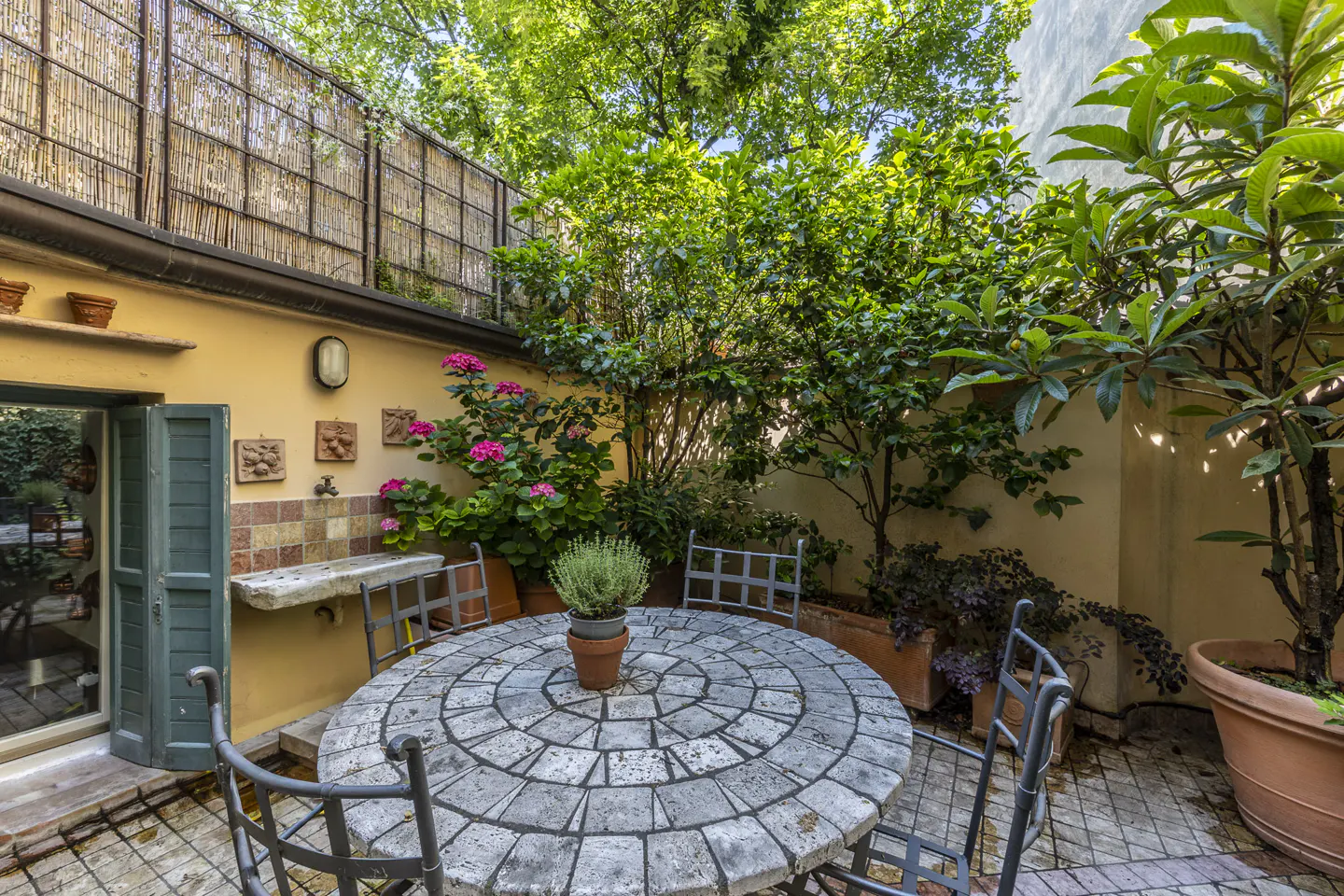 Outdoor patio with stone table, metal chairs, and lush greenery. A yellow wall with a sink and decorations adds charm.