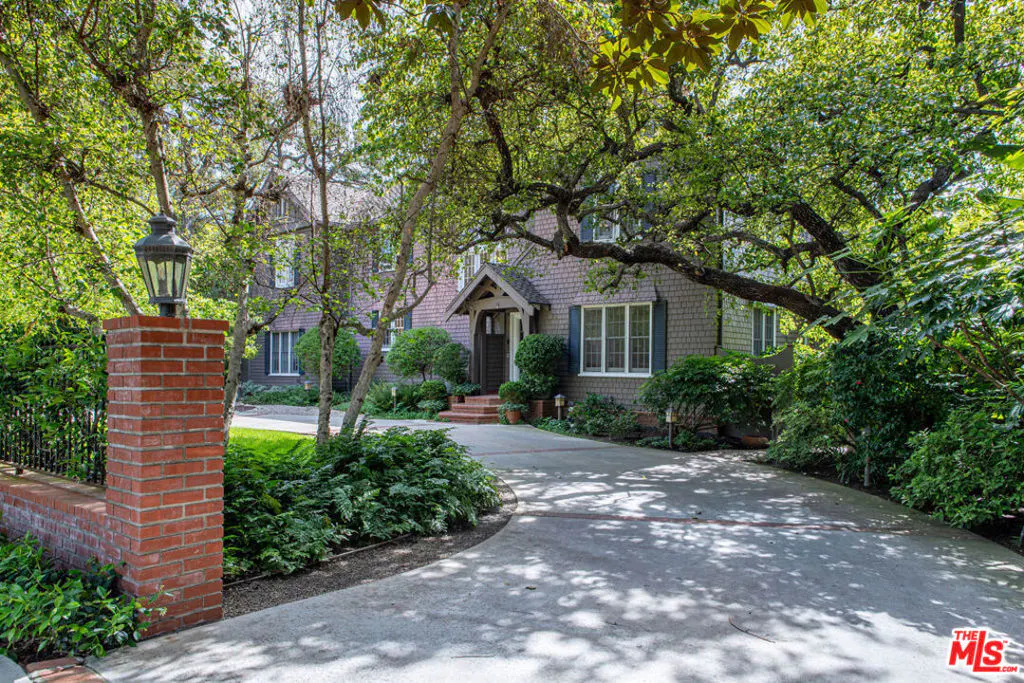 Exterior view of a two-story house with gray shingles, green shutters, and a concrete driveway lined with trees and greenery.