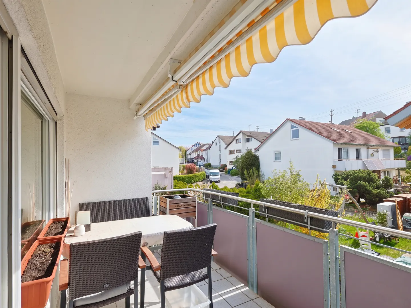 Balcony with a table, chairs, and a bench. A yellow and white striped awning provides shade. Houses are visible in the background.