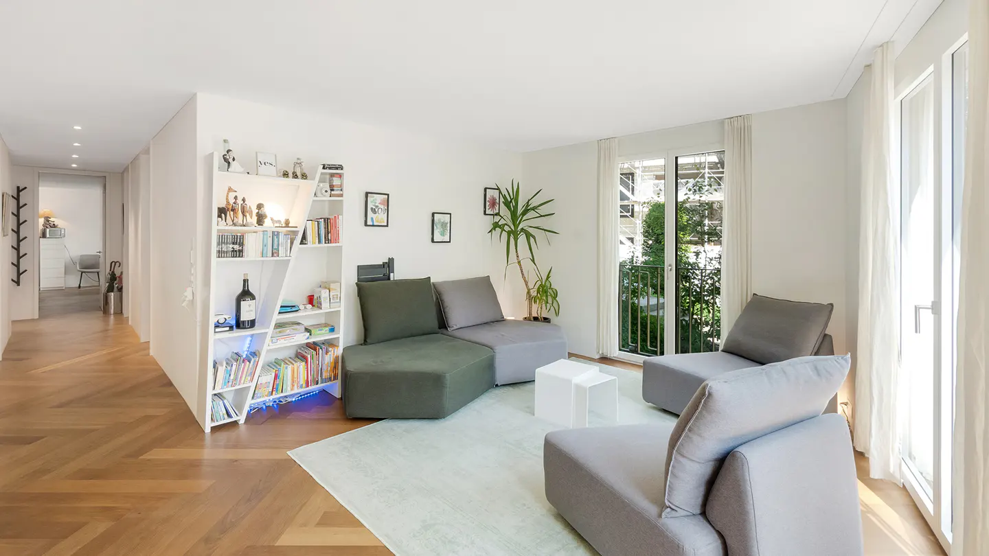 Bright living room with modular gray and green sofa, white bookshelf, and balcony doors with sheer curtains.