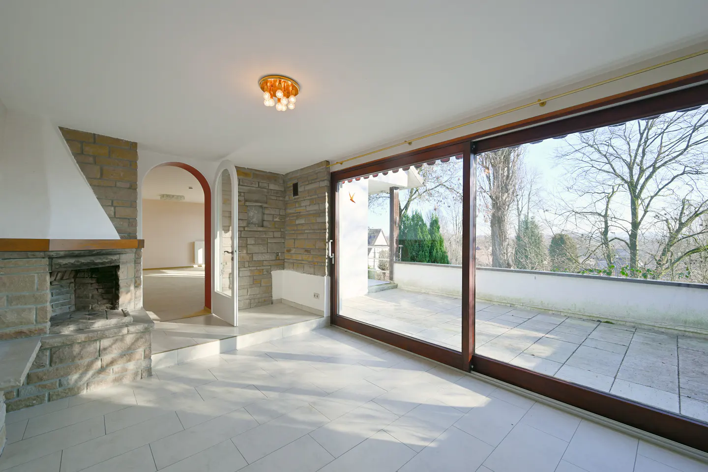 Bright living room with stone fireplace, white tile floor, and large sliding glass doors to a patio with trees in the background.