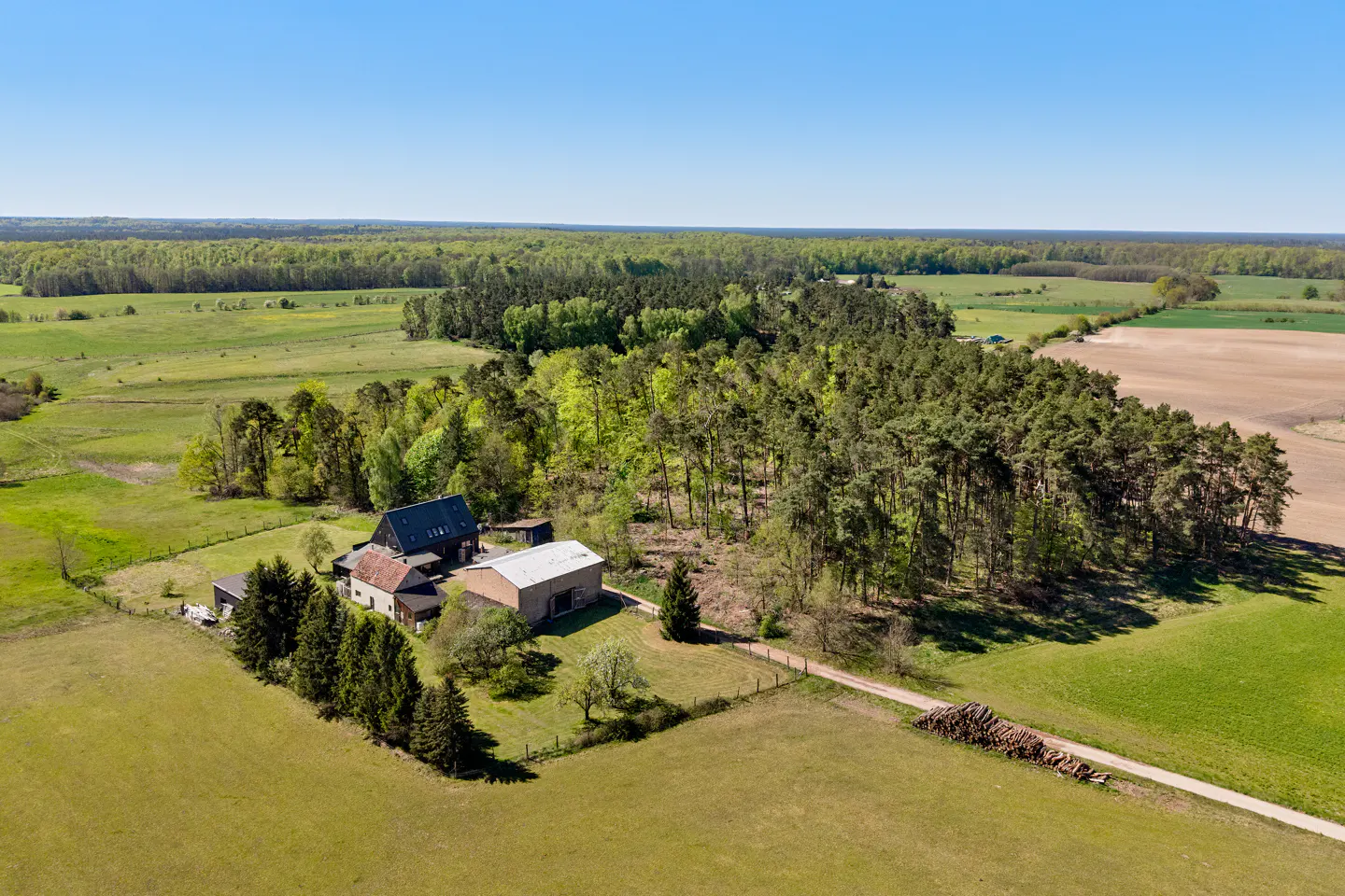 Aerial view of a rural property with a house, barn, and surrounding green fields and trees under a clear blue sky.
