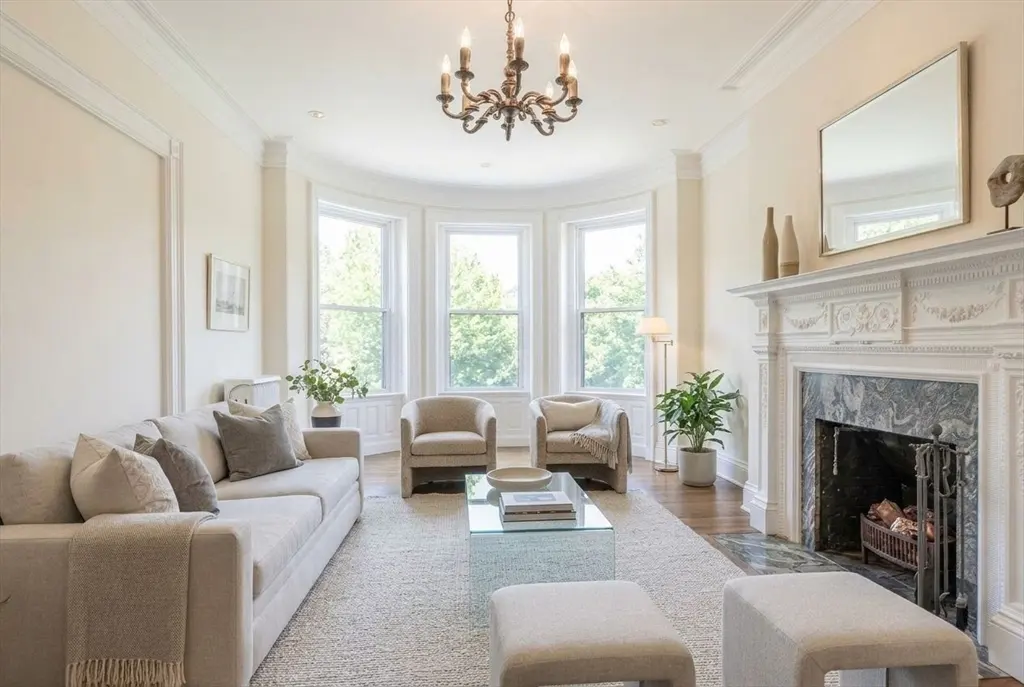 Bright living room with beige sofa, chairs, and rug. A fireplace with a white mantel and a chandelier add elegance. Large windows offer a view of green trees.