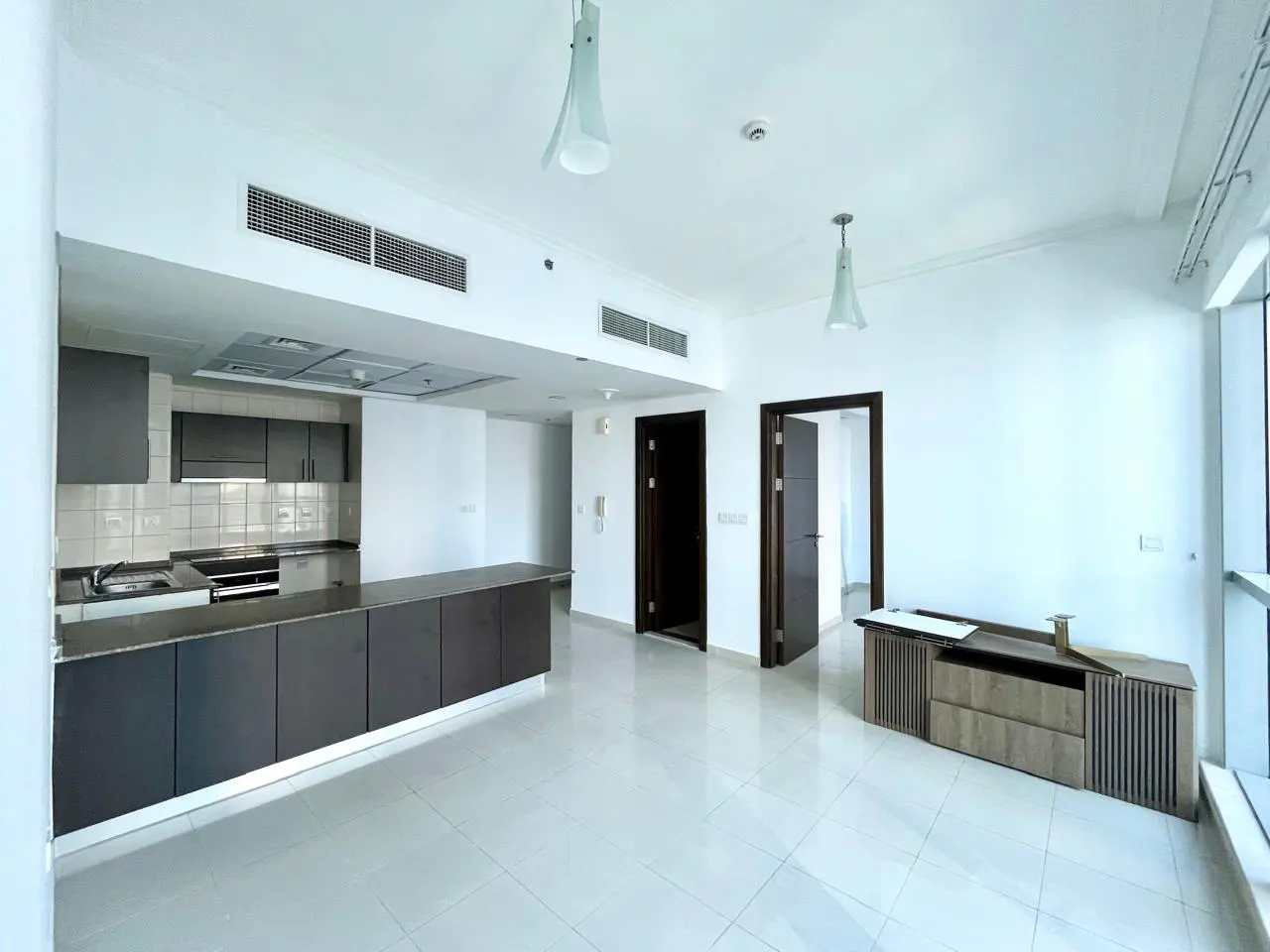 Bright, modern apartment interior with gray kitchen cabinets, white walls, and tile floors. A wooden TV stand sits near a window.