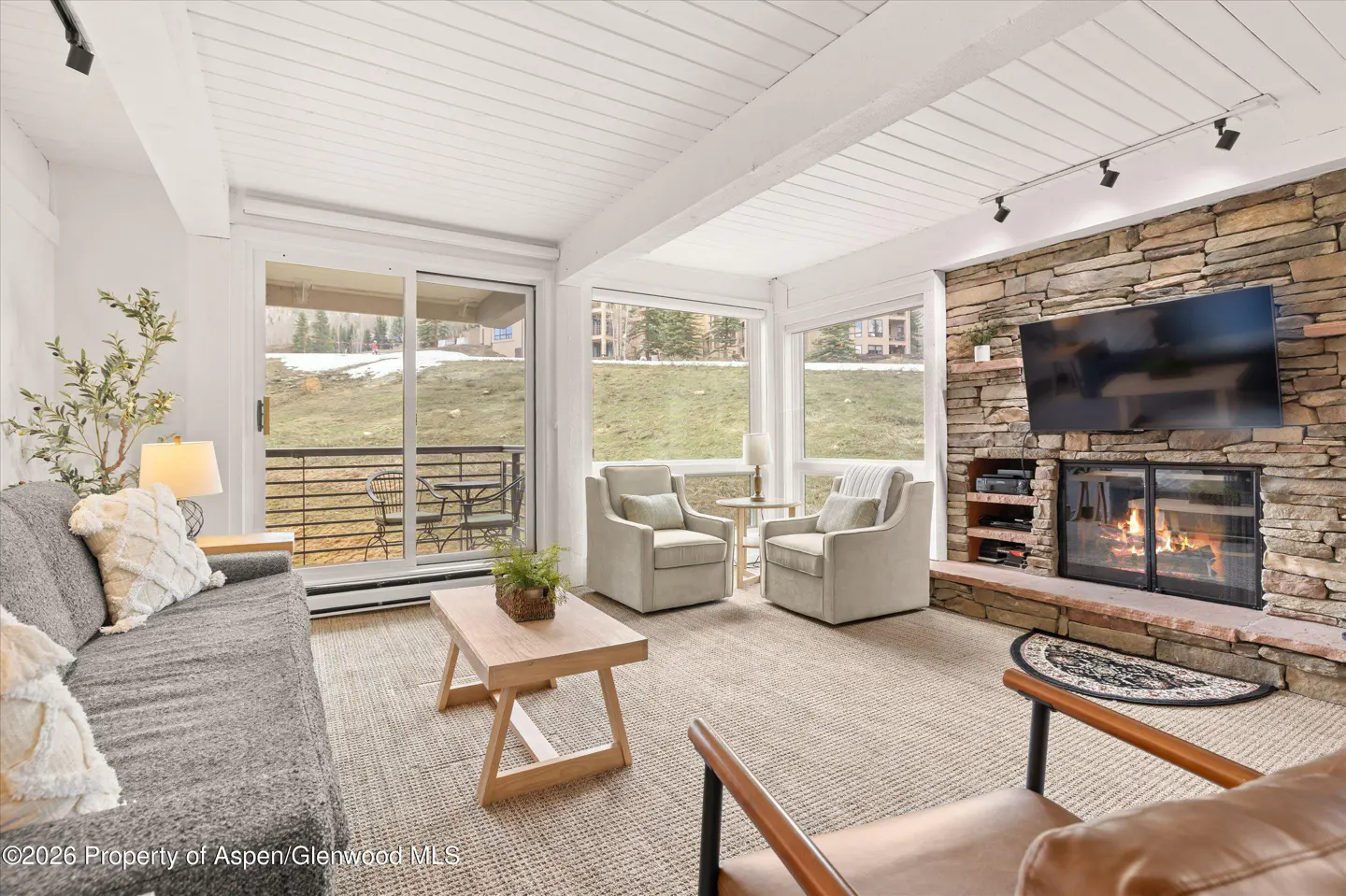 Living room with a stone fireplace, TV, gray sofa, two armchairs, and a wooden coffee table. Large windows overlook a grassy hill.