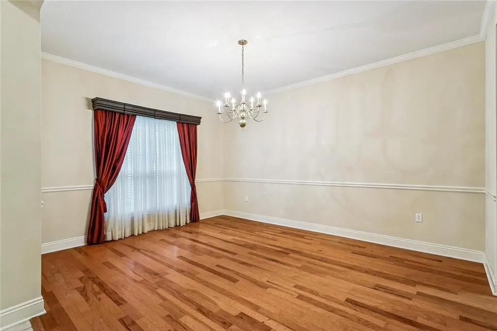 Empty dining room with hardwood floors, beige walls, and a chandelier. Red curtains frame a window.