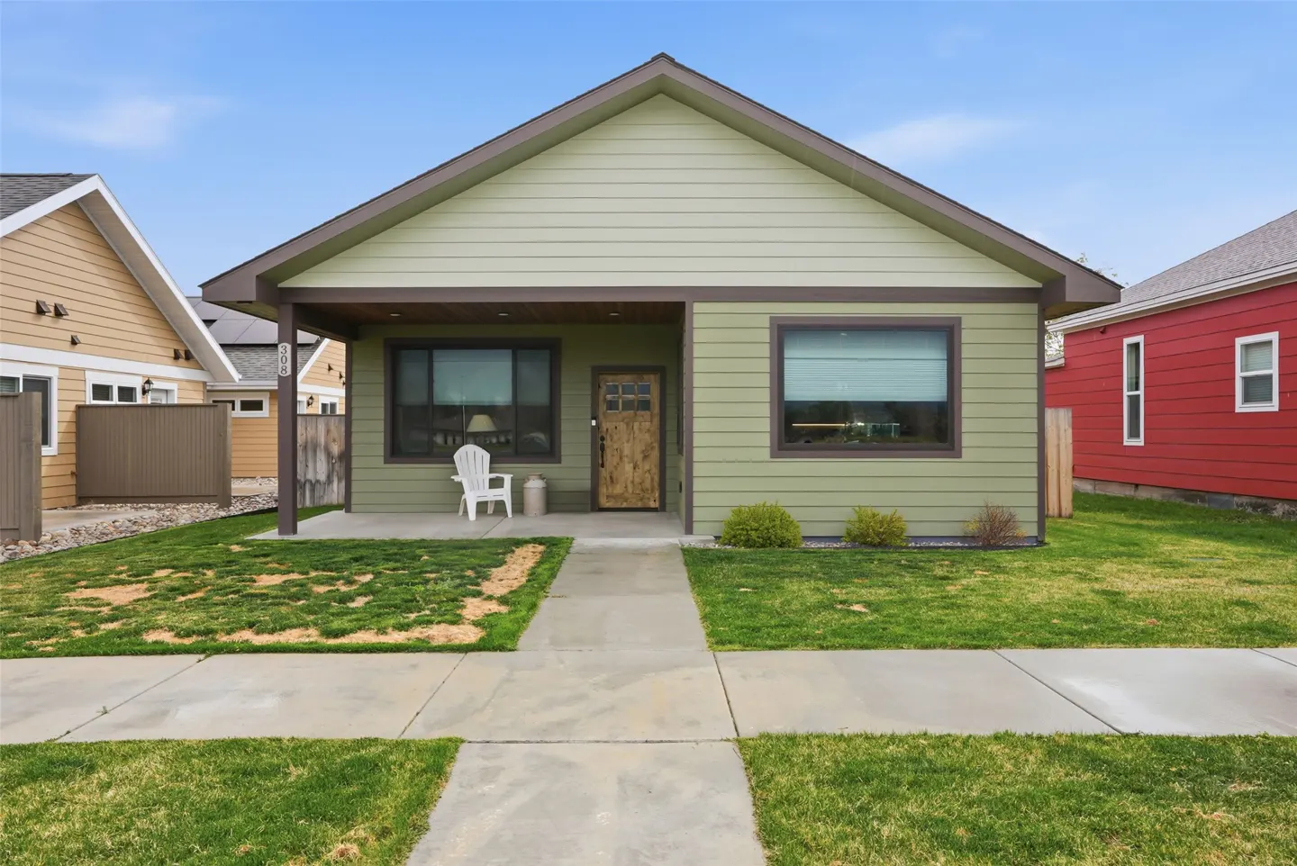 Front view of a one-story, light green house with a brown-trimmed porch, a wooden door, and a white chair on the porch.