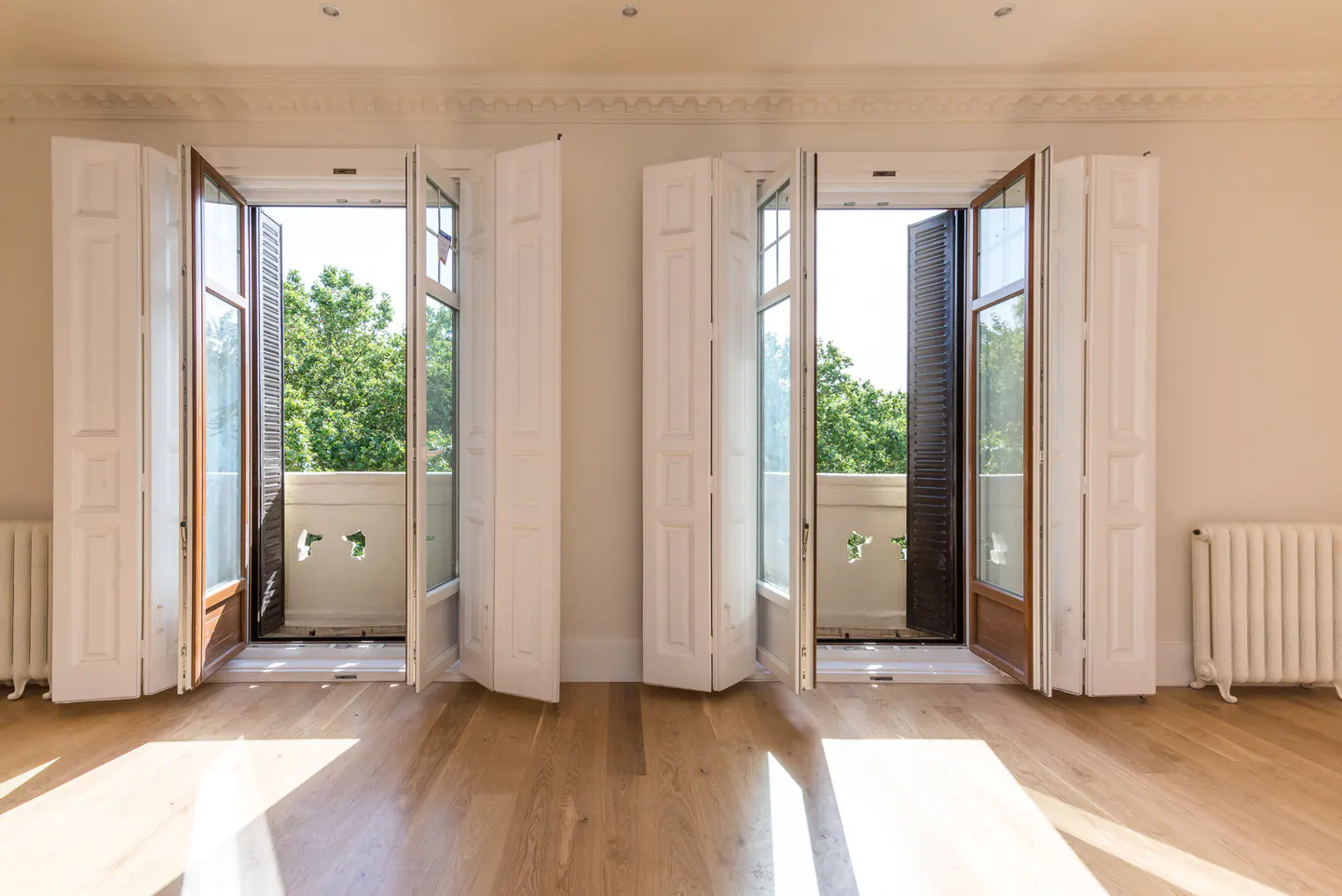 Two open white French doors with wooden shutters reveal a balcony with green trees against a light-filled room.