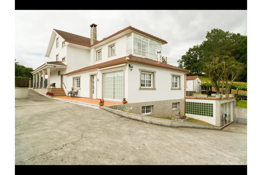 A white multi-story house with a terracotta roof, stone foundation, and a concrete driveway under a cloudy sky.