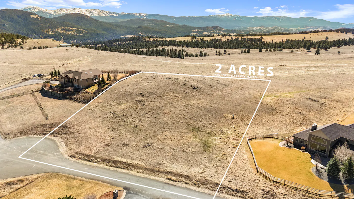 Aerial view of a 2-acre vacant lot outlined in white, with mountains, trees, and houses in the background.