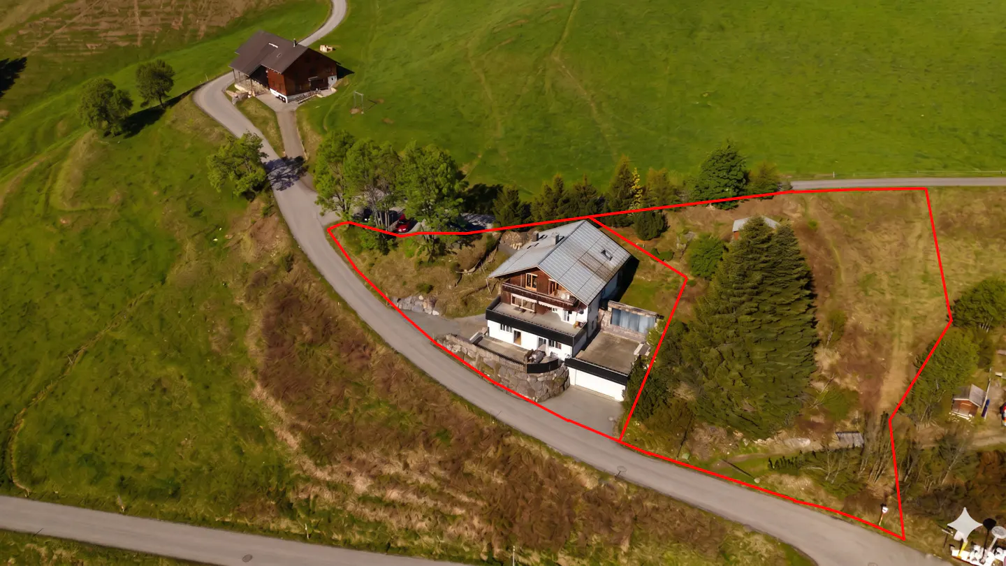 Aerial view of a two-story house with a gray roof, surrounded by a red boundary line, on a green hillside.