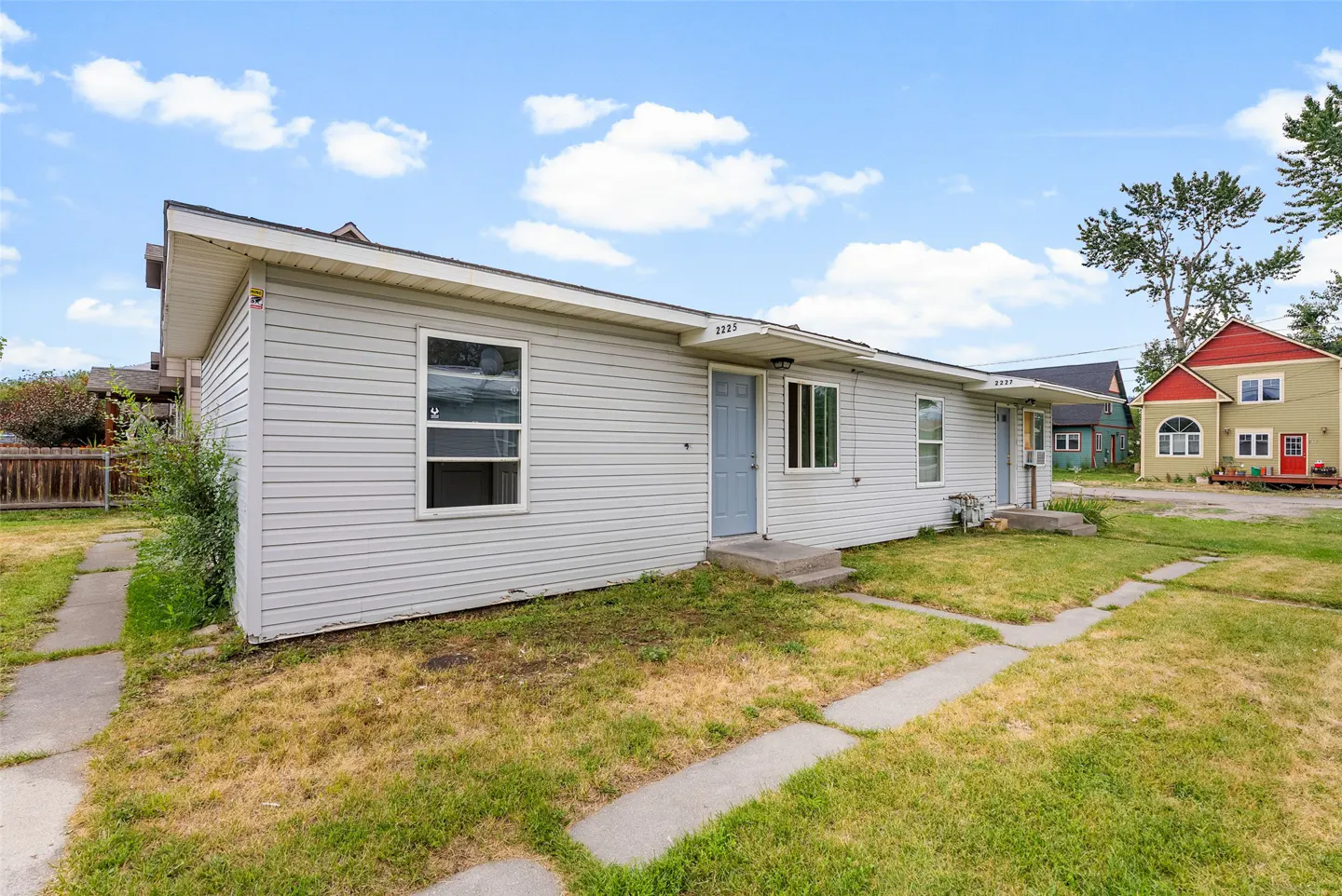 A light gray duplex with blue doors and white trim, set on a lawn with a concrete walkway. A colorful house is visible in the background.