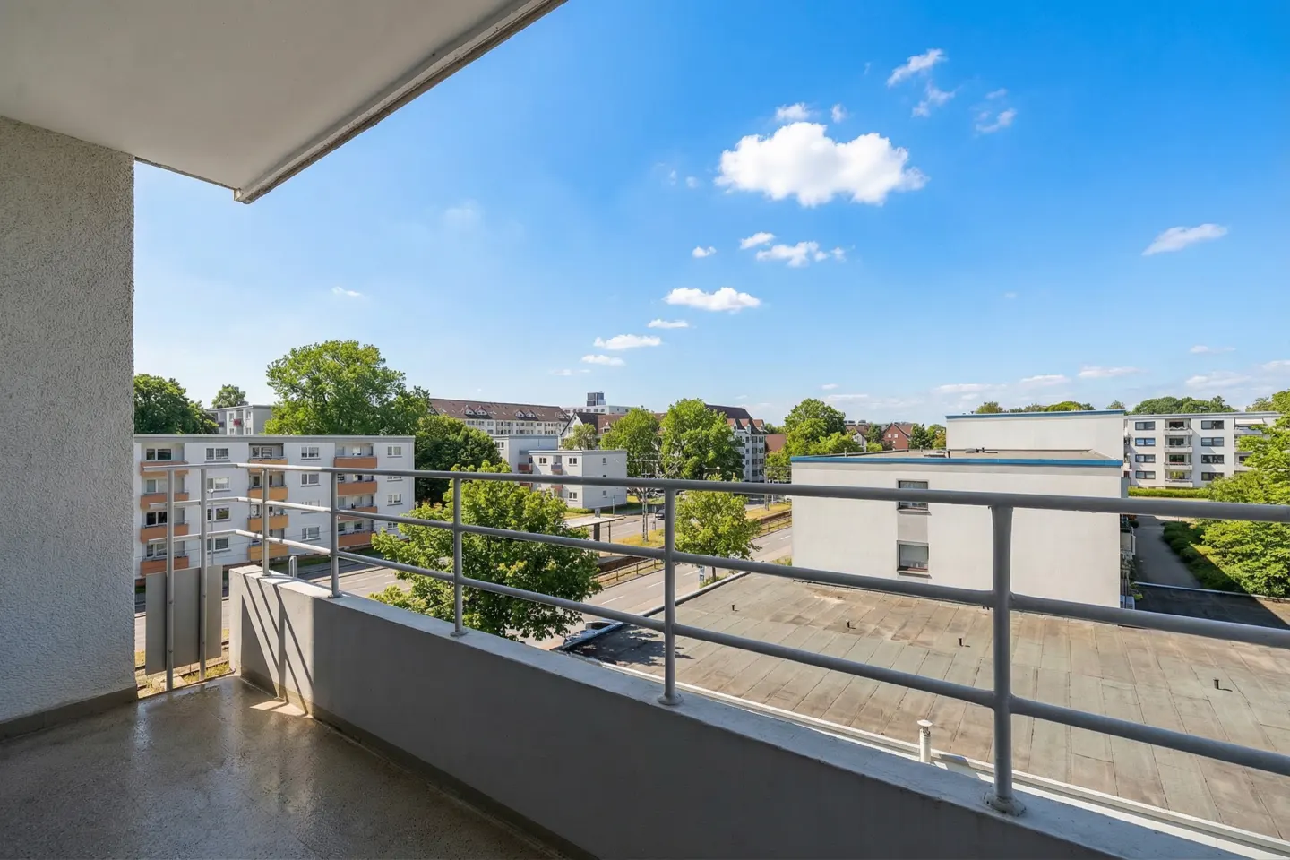 View from a balcony with gray railings overlooking buildings, trees, and a blue sky with white clouds.