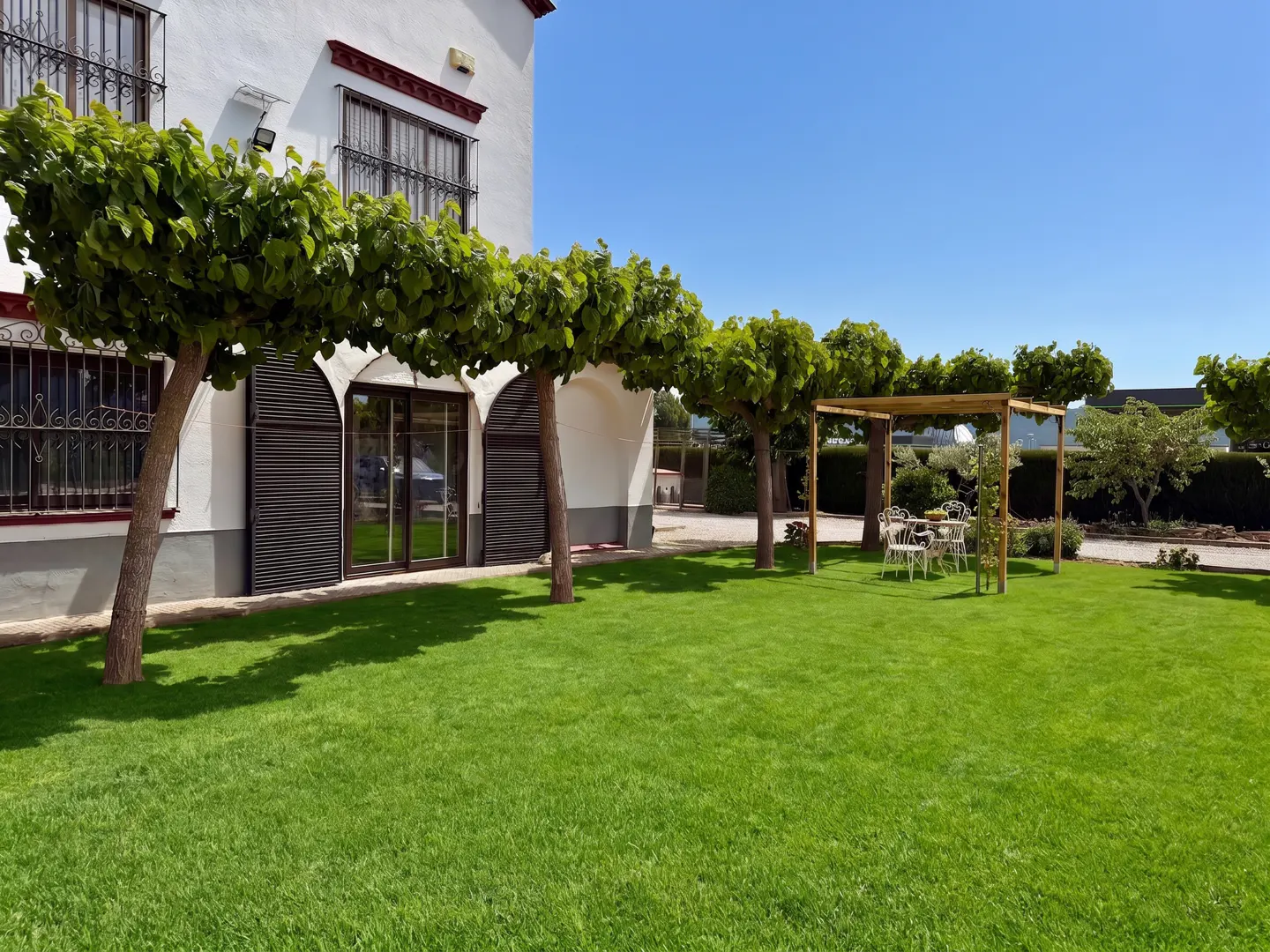 Exterior of a white building with a green lawn, trees, and a pergola with a table and chairs.