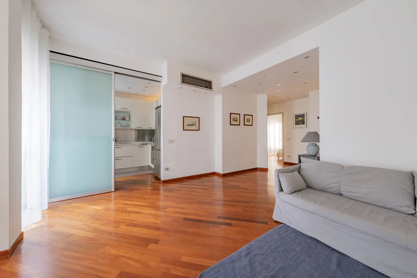 Bright, open-concept living space with hardwood floors, white walls, and a gray sofa. A sliding glass door reveals a modern white kitchen.