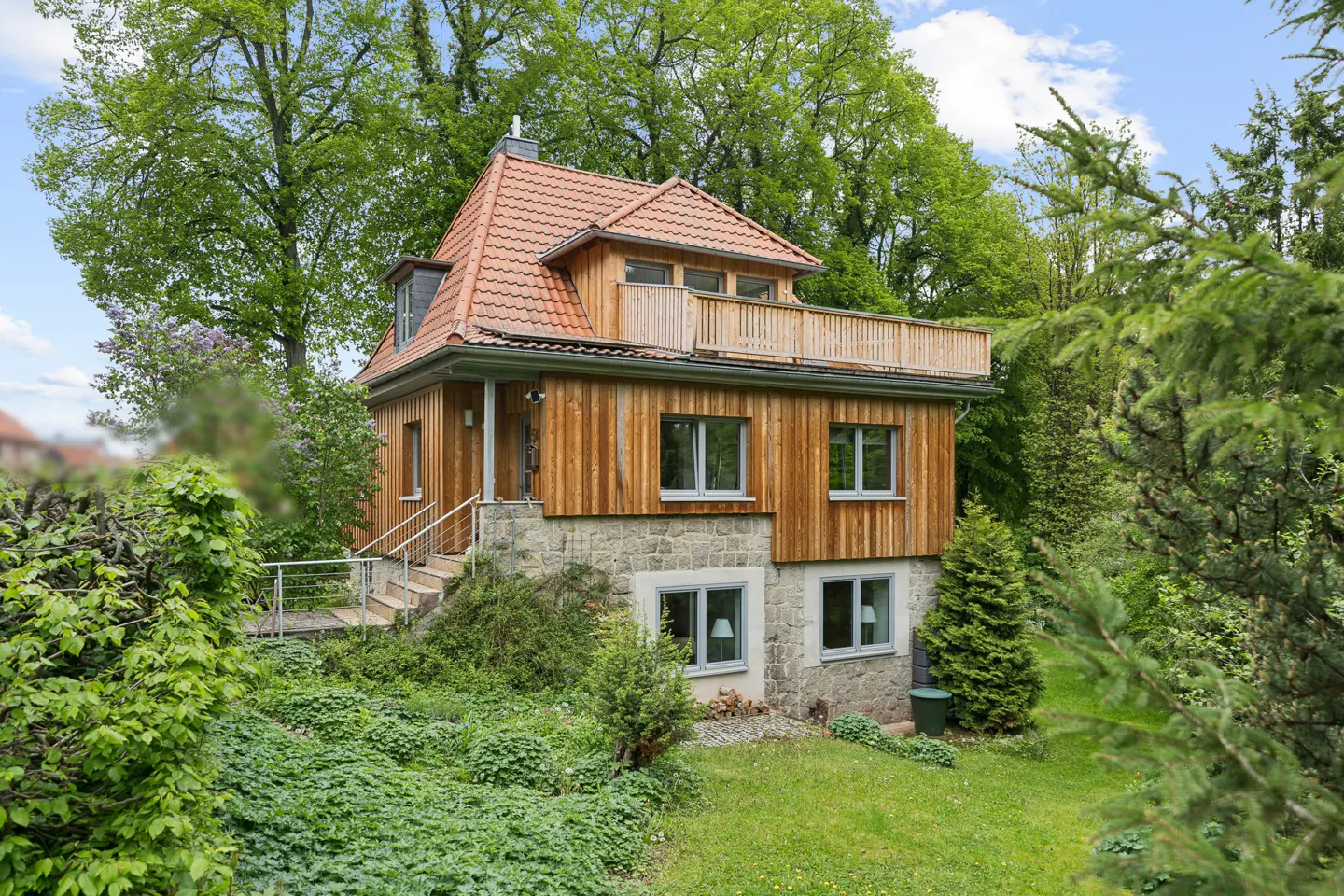 Two-story house with wood siding, stone foundation, and red tile roof, surrounded by lush greenery.