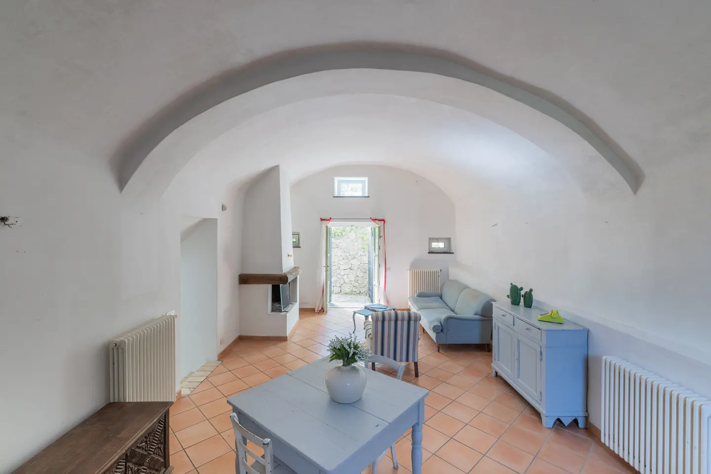 Bright living room with arched ceiling, terracotta tile floor, fireplace, and blue furniture. Open doorway leads to a stone patio.