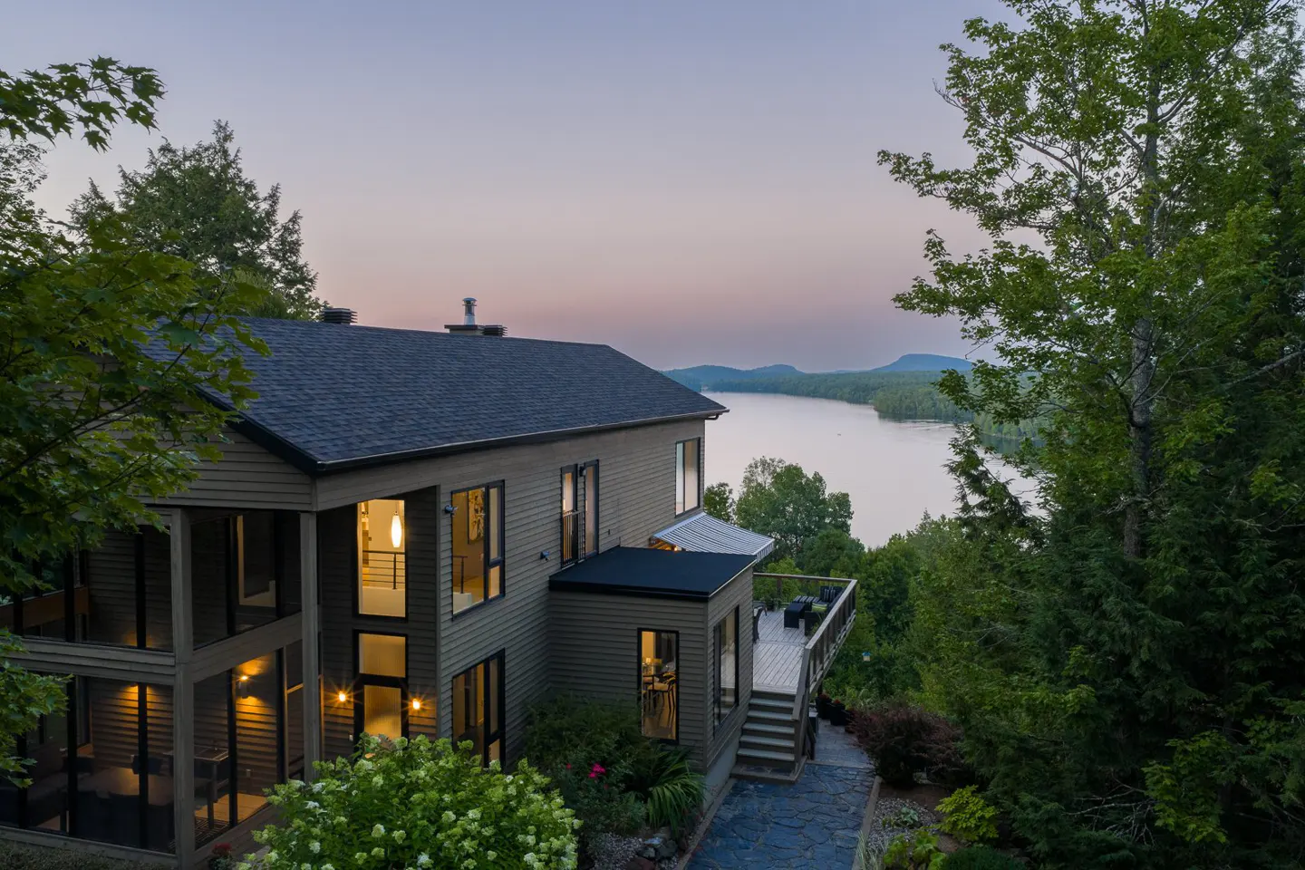 Exterior view of a modern, two-story house with a dark roof, overlooking a lake at dusk, surrounded by green trees.