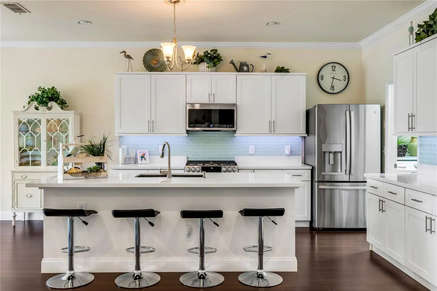 Bright kitchen with white cabinets, stainless steel appliances, and a large island with four black bar stools. Dark wood floors and light yellow walls.