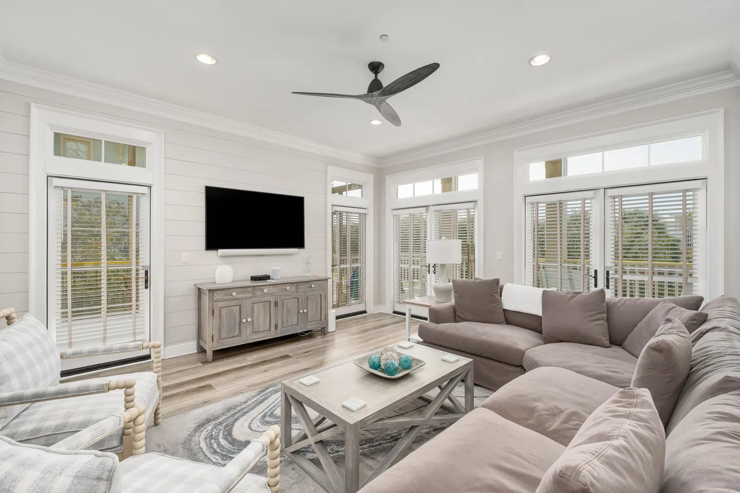 Bright living room with a gray sectional sofa, TV, and sliding glass doors. A ceiling fan hangs above a wooden coffee table.