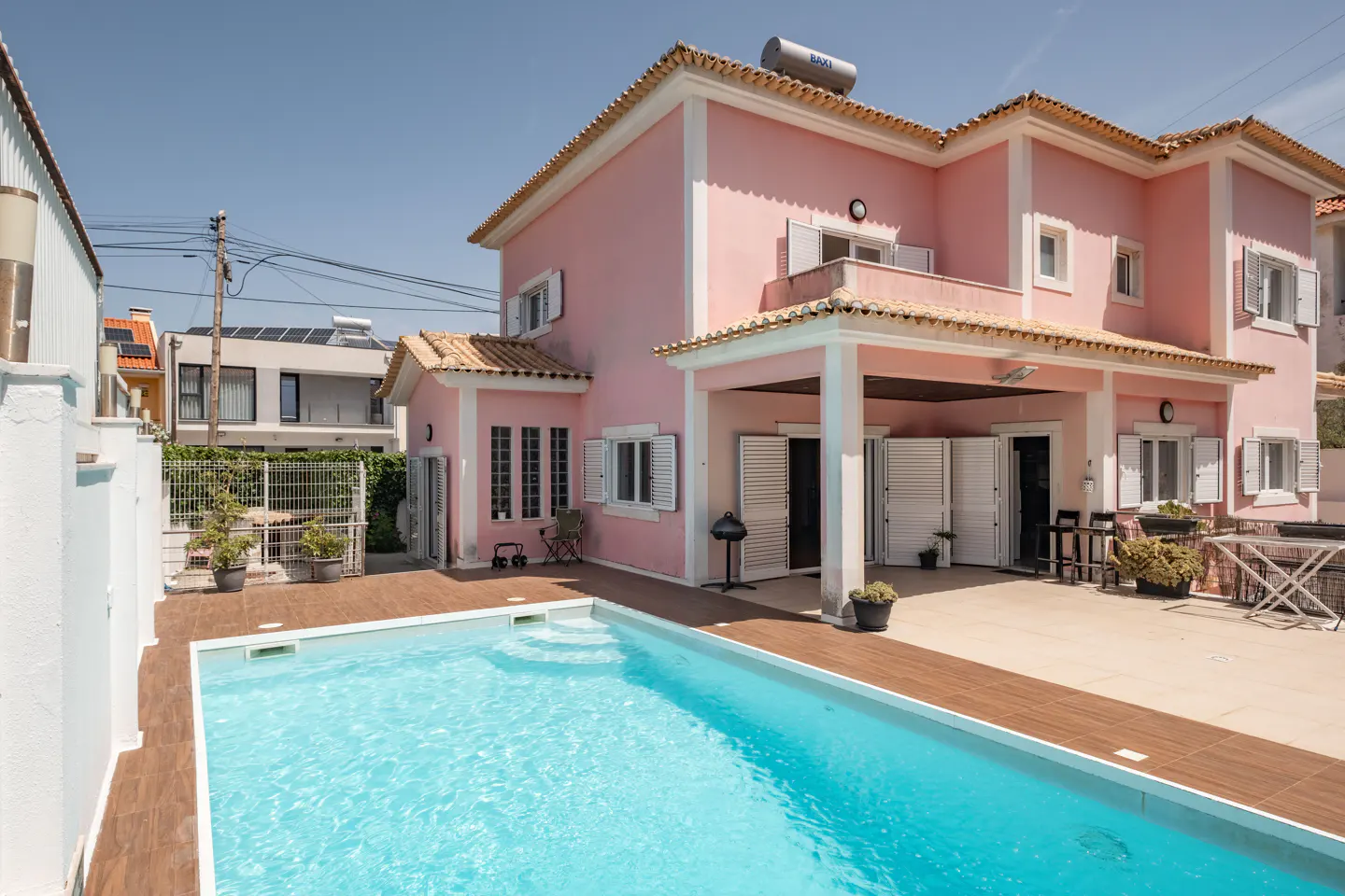 A two-story pink house with a pool and a brown tiled patio on a sunny day.