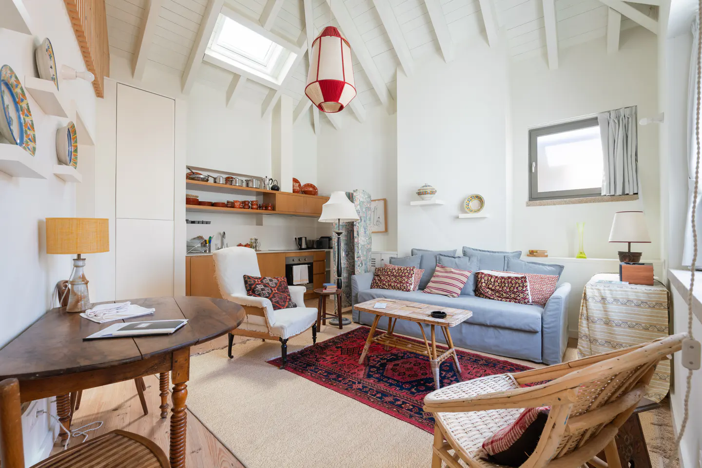 Bright, airy living space with white walls, a blue sofa, and a red patterned rug. A small kitchen area is visible in the background.