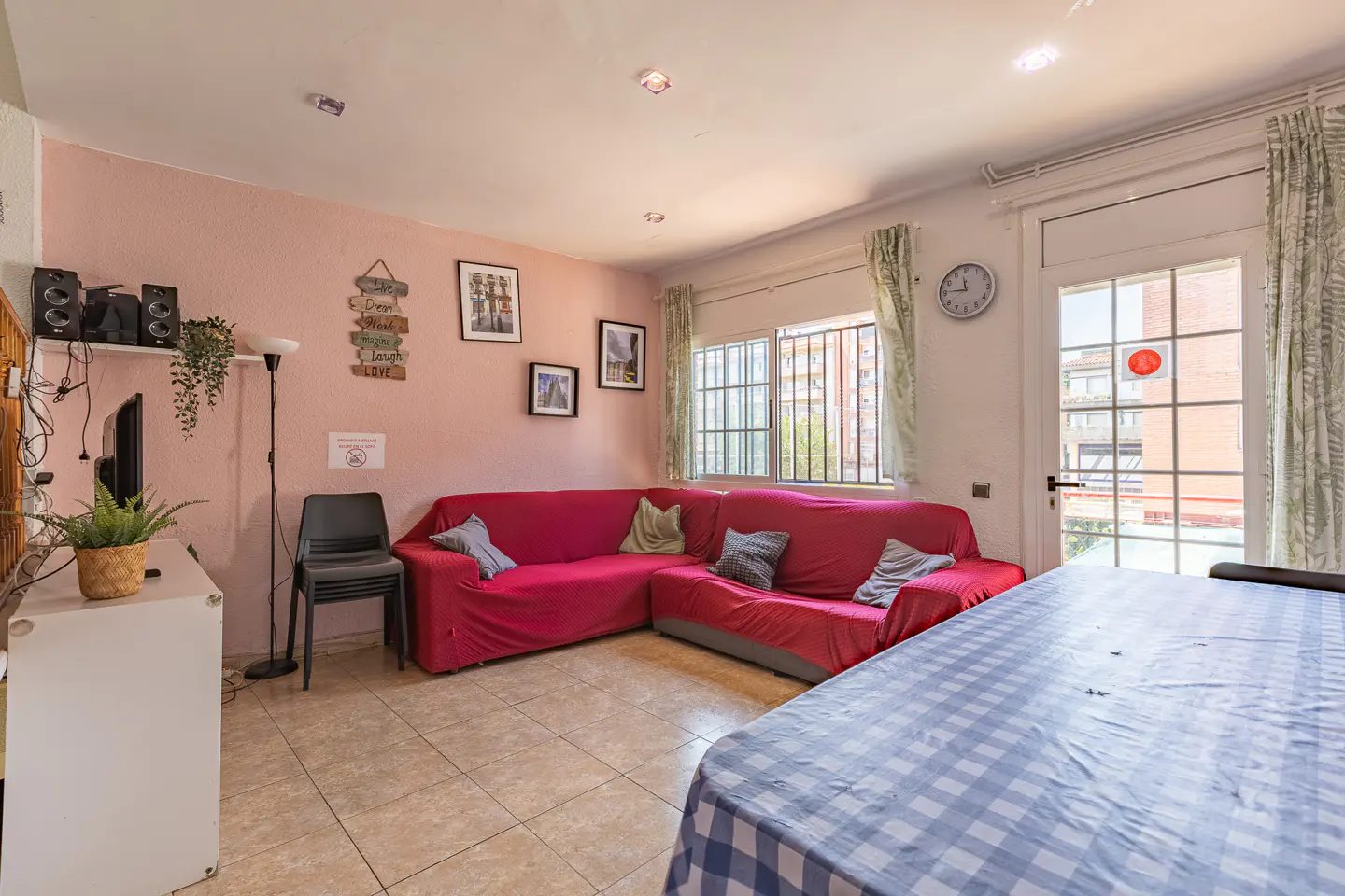 Living room with pink walls, a red L-shaped sofa, a dining table with a blue checkered cloth, and a glass door to a balcony.