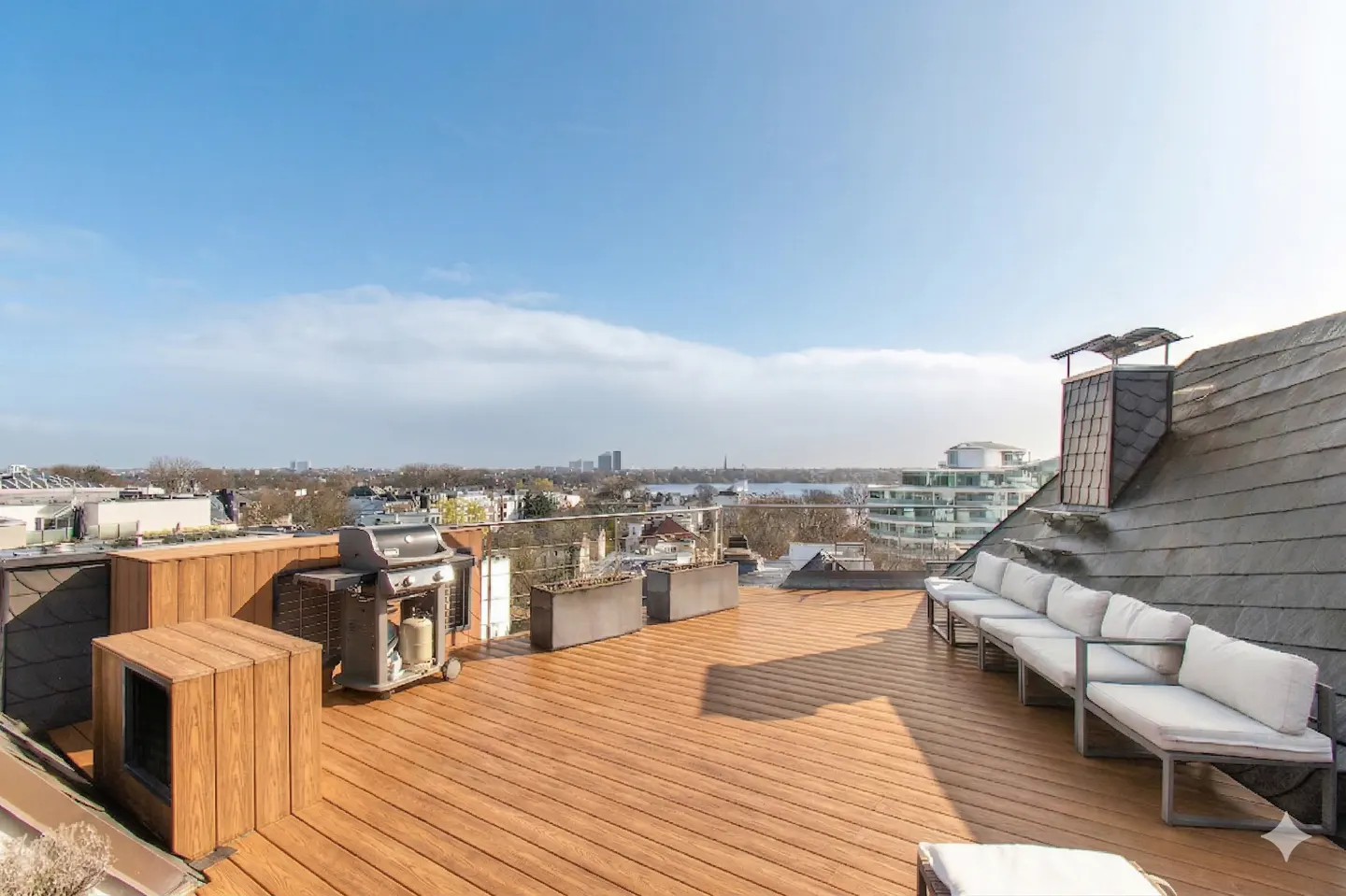 Rooftop patio with wood deck, white cushioned seating, grill, and city view under a blue sky.