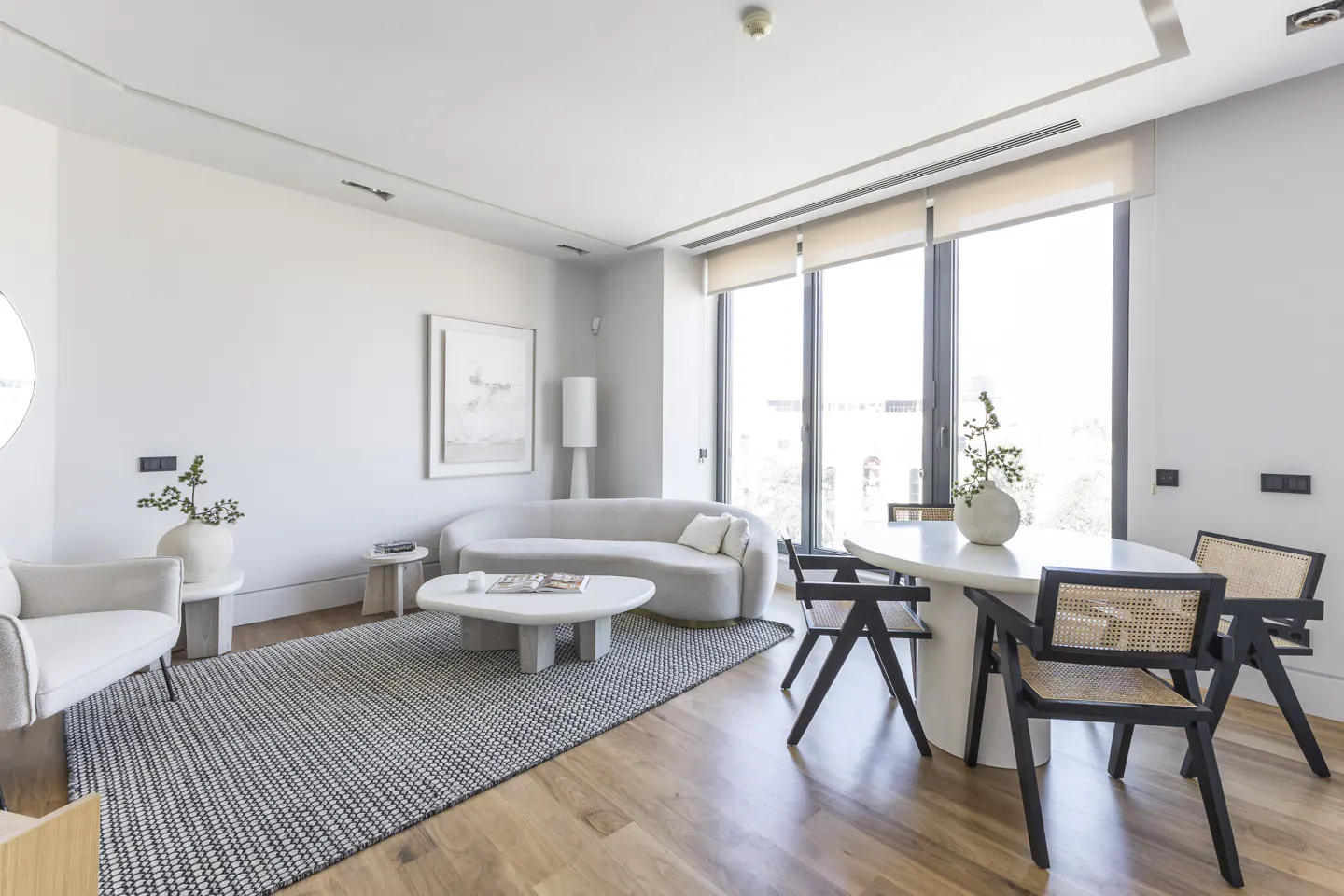 Bright, modern living room with a curved sofa, stone coffee table, and dining area with black chairs. Wood floors and large windows.