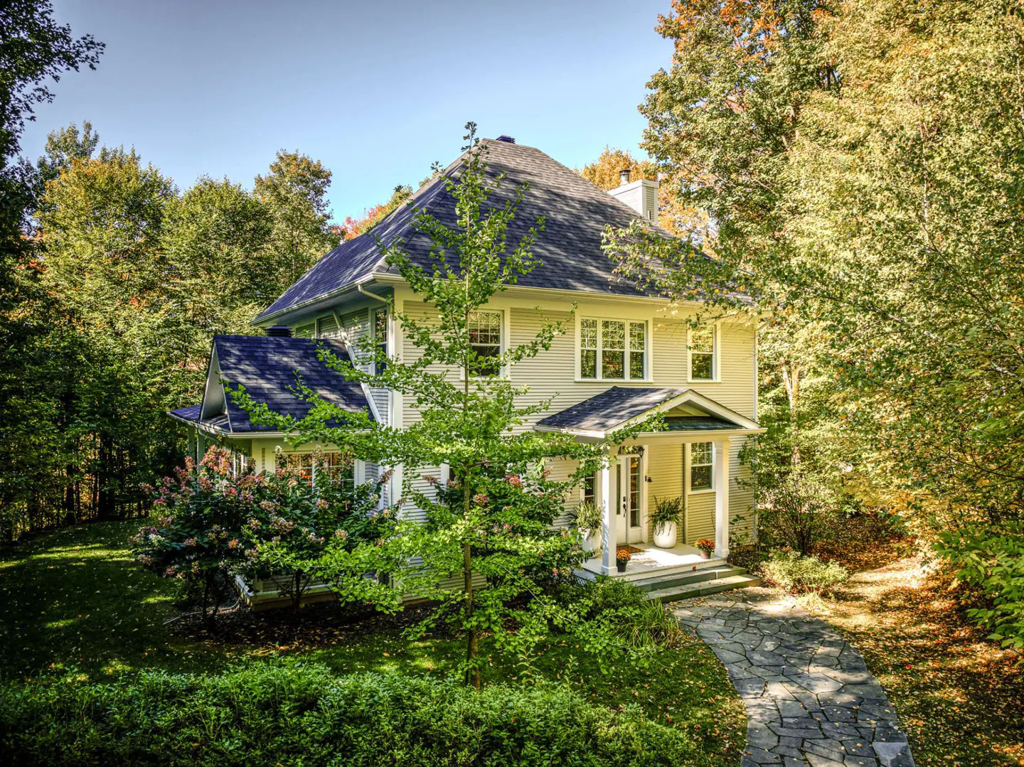 Two-story light yellow house with a dark roof, surrounded by green trees and bushes. A stone path leads to the front door.