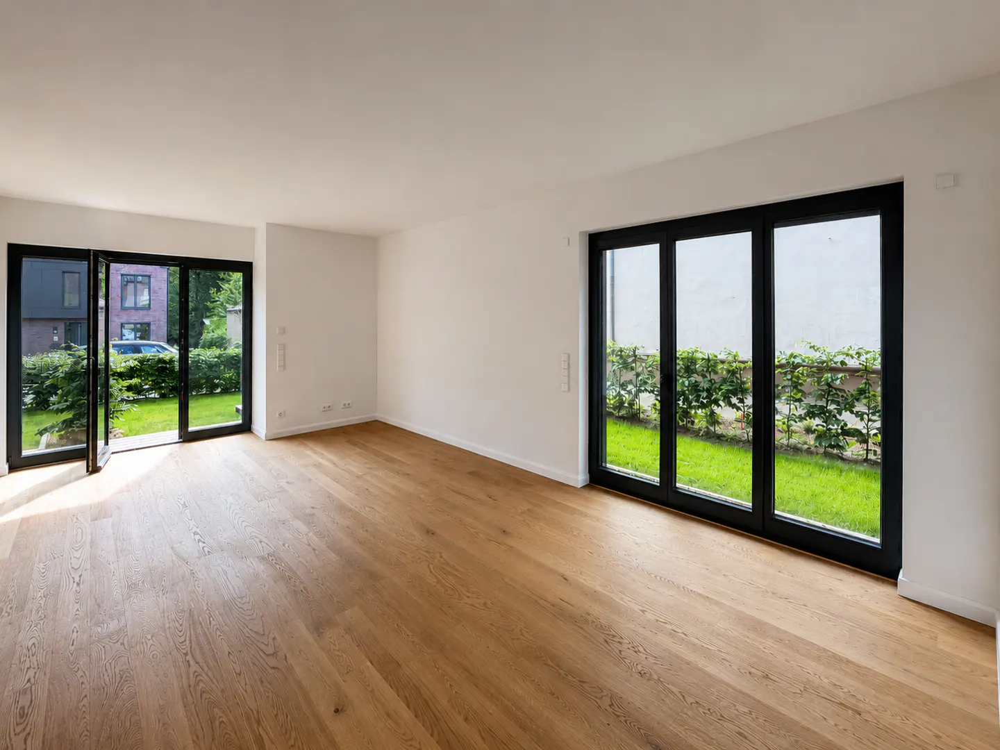 Empty room with light wood floors, white walls, and large black-framed glass doors opening to a green garden.