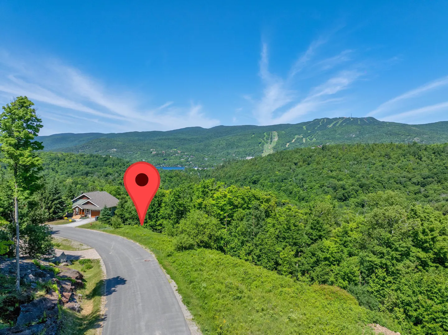 Scenic view of a property with a red map pin, a house, a road, and a mountain range in the background under a blue sky.