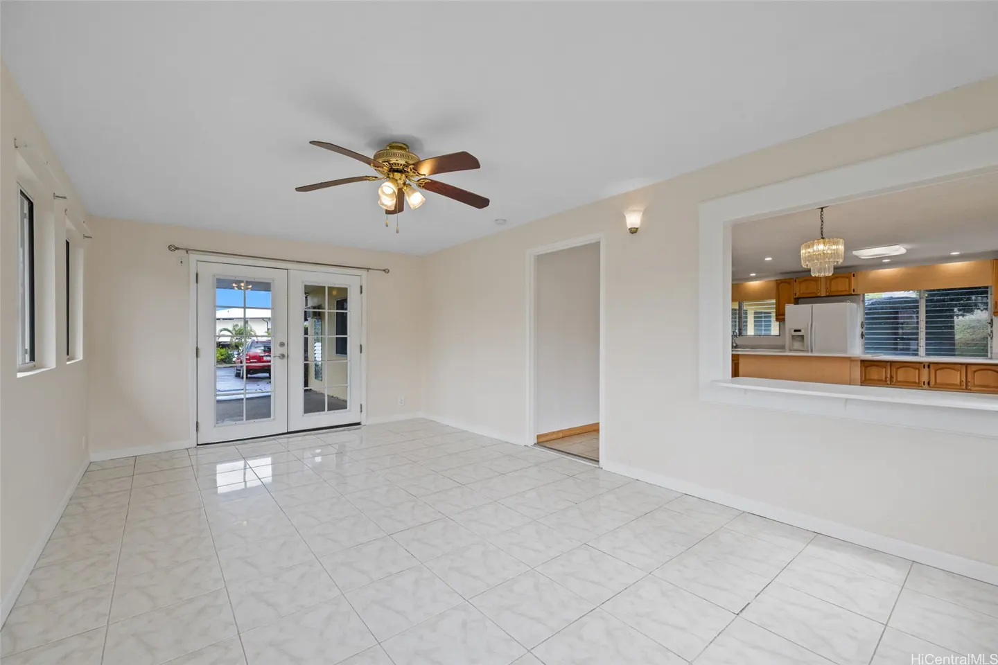 Bright, empty room with white tile floor, ceiling fan, and French doors. A kitchen is visible through a window.