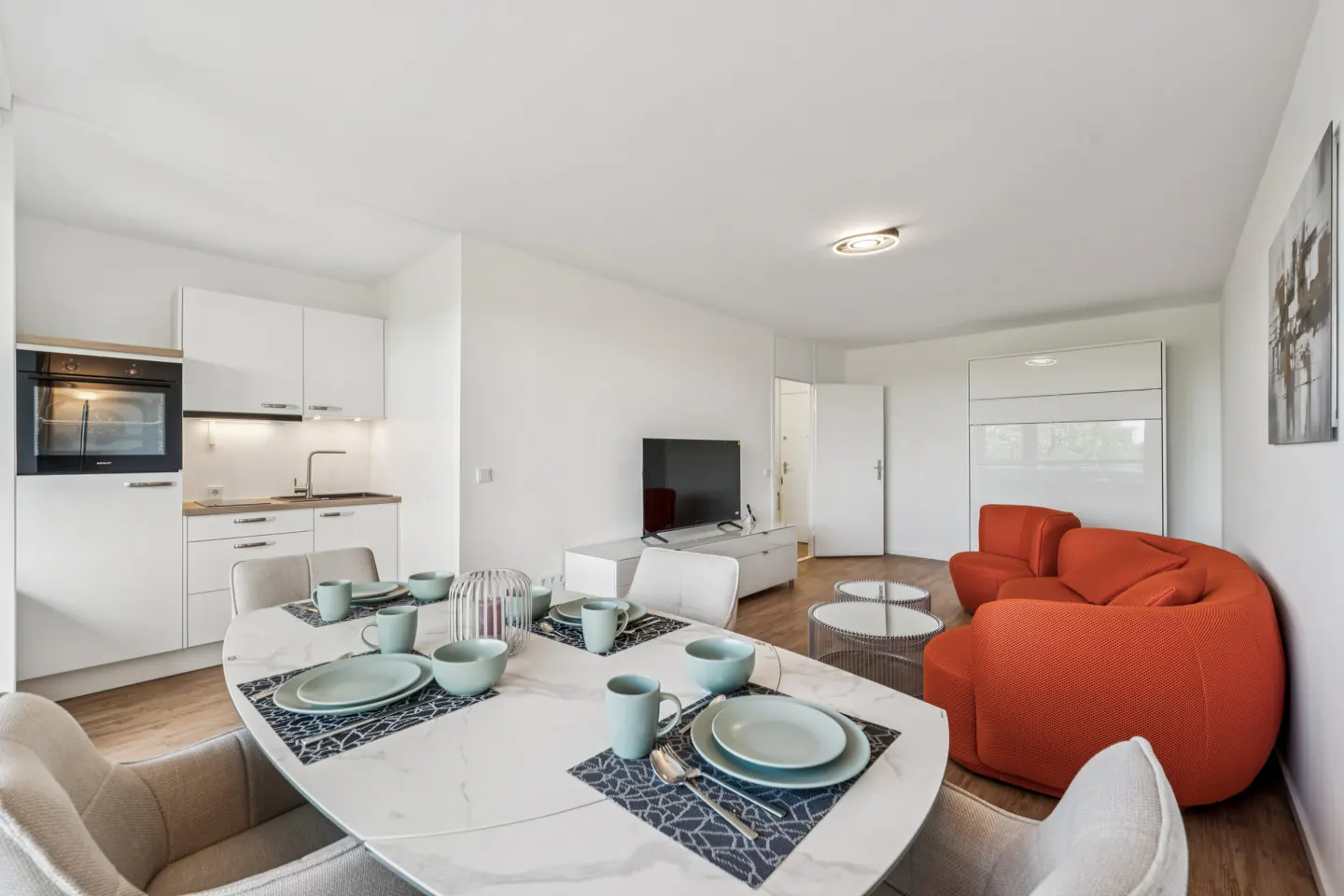 Bright, modern apartment with white walls, kitchen, and orange sofa. Dining table set with blue dishes and gray chairs.
