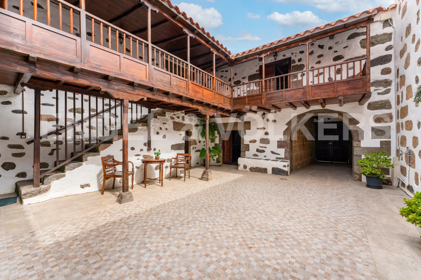 Courtyard view of a two-story white building with dark stone accents and wooden balconies. A table and chairs sit on the tiled floor.