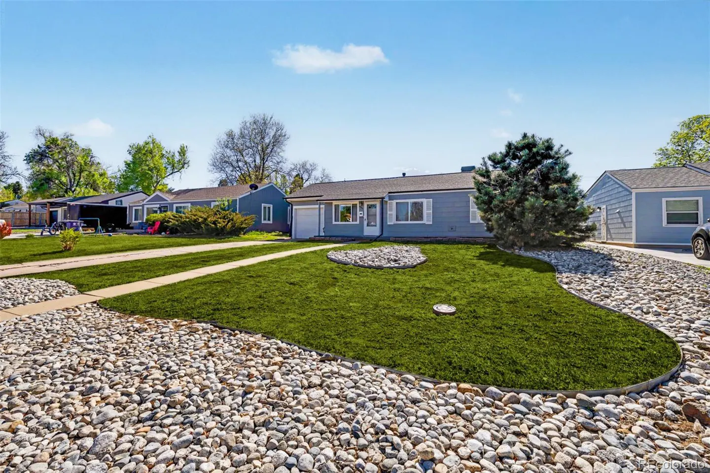 A light blue single-story house with a green lawn and rock landscaping under a blue sky.