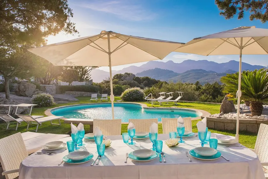 Outdoor dining table set with turquoise glasses and plates, white tablecloth, pool, and mountain view.