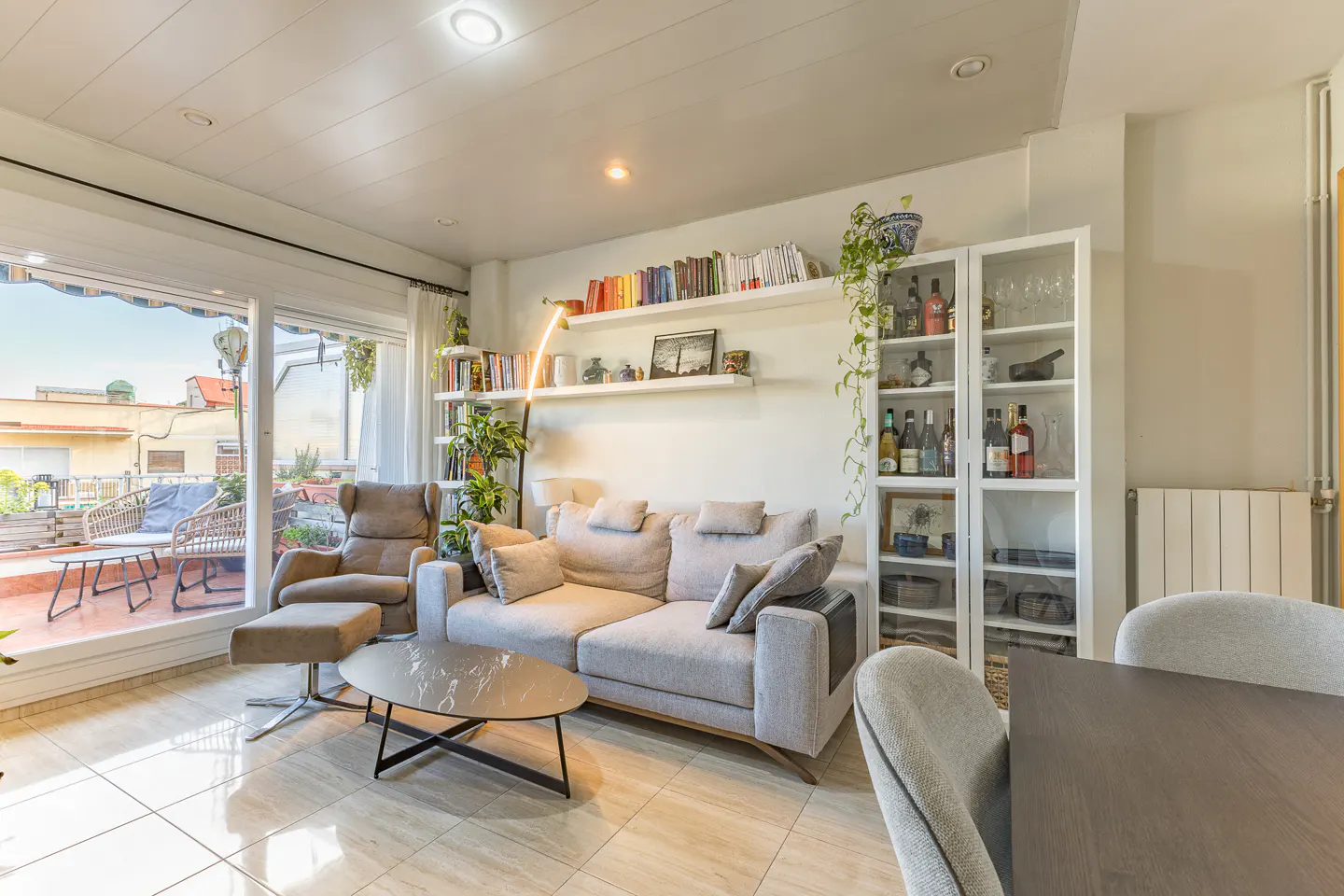 Bright living room with a gray sofa, chair, and coffee table. Bookshelves and a cabinet line the wall. A balcony with outdoor furniture is visible.