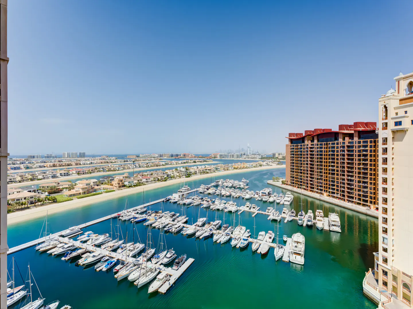 Aerial view of a marina filled with white yachts, next to a brown building, under a clear blue sky.