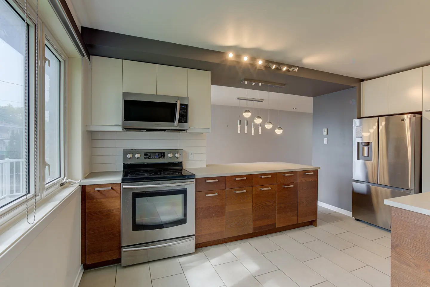 A modern kitchen with stainless steel appliances, wood cabinets, and white countertops. A large window provides natural light.